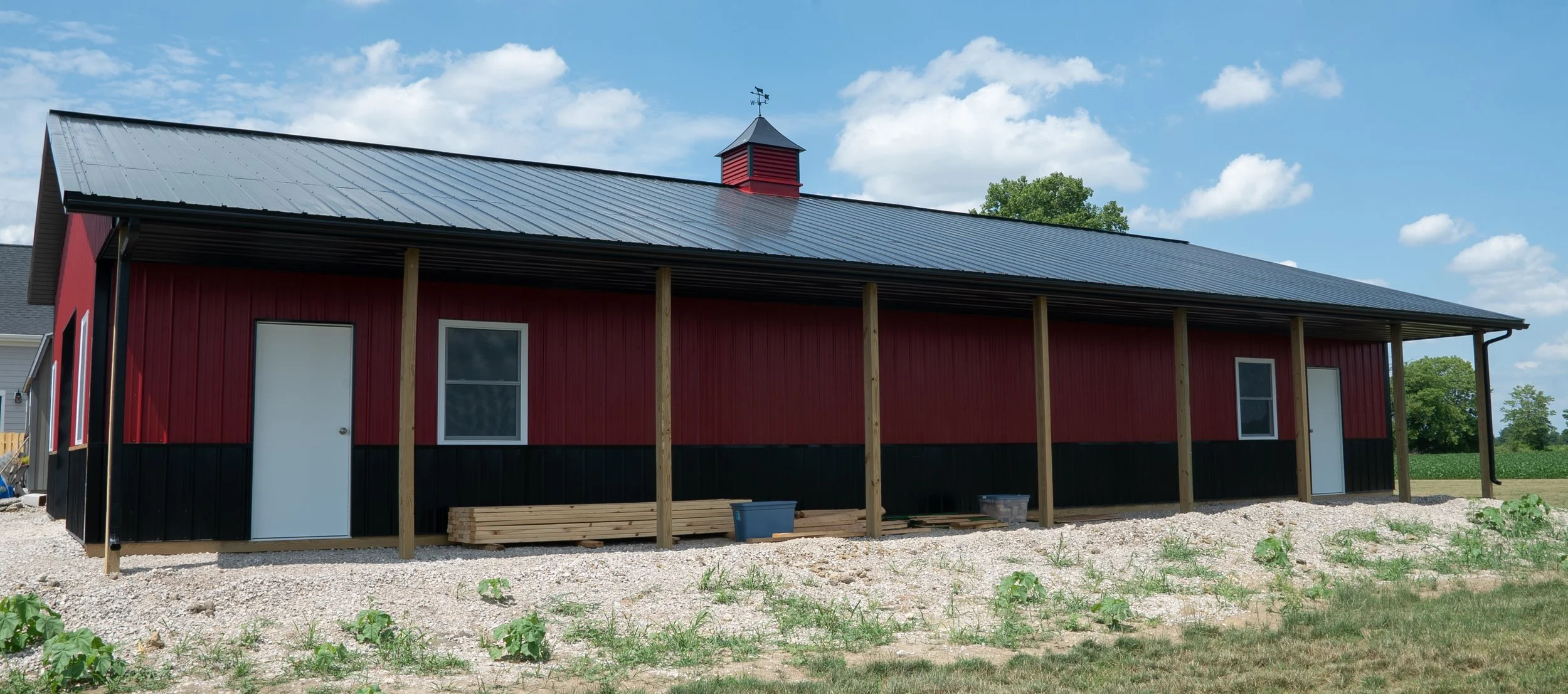 A red barn with black lower siding, white doors, and windows. It has a metal roof and a small cupola on top with a weather vane. Wooden posts are positioned along the front, and construction materials are on the ground nearby. The background features