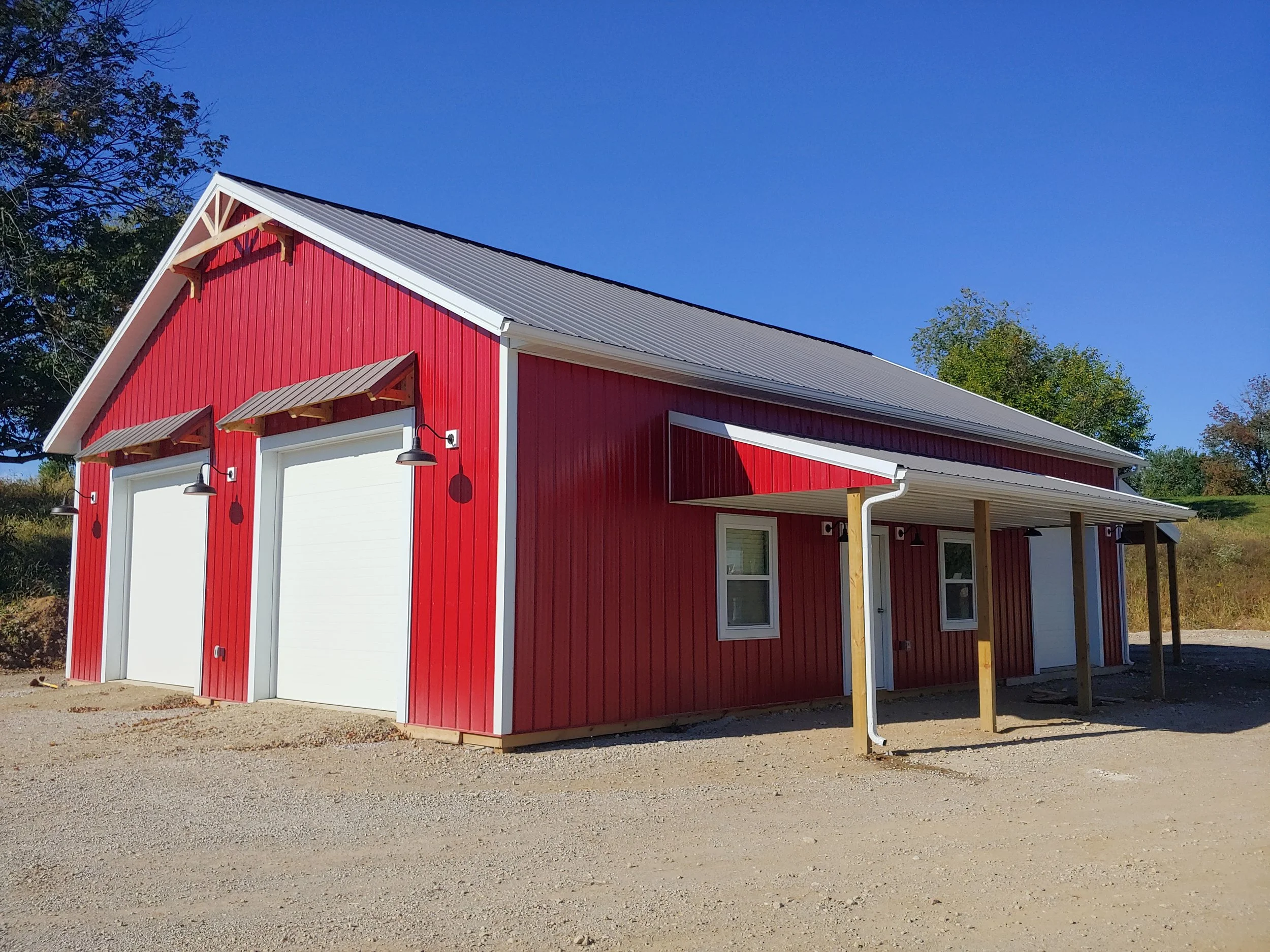A red barn-style building with white trim, garage doors, and outdoor wall-mounted lights, set against a clear blue sky with trees in the background.