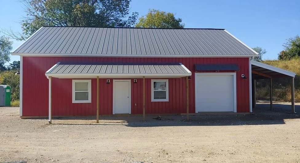 A red barn-style building with a metal roof, two windows, a central door, and a garage door. There is a small covered porch in front, supported by wooden posts, and a carport on the right side.