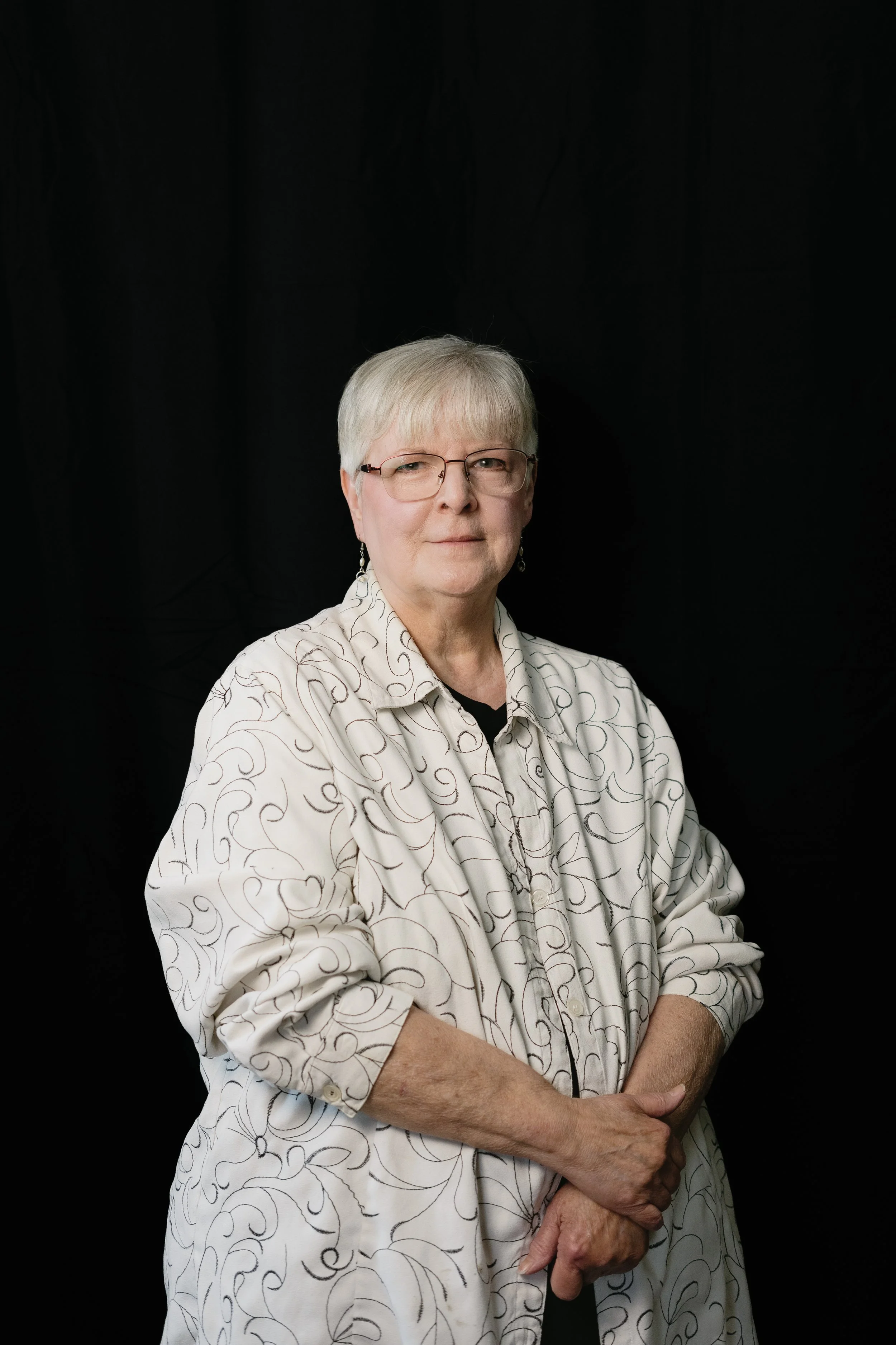 An elderly woman with short gray hair, wearing glasses and a light-colored blouse with a swirl pattern, standing against a black background.
