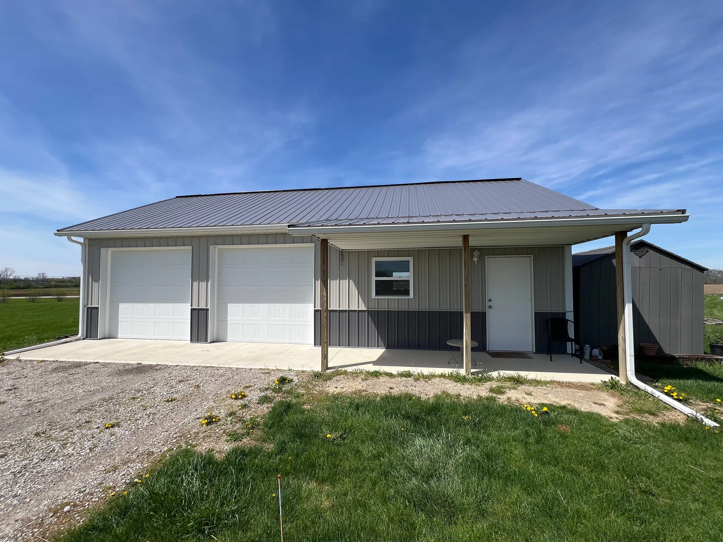 A house with a metal roof, white siding, a garage door, and a small porch with supports. The yard has green grass and small yellow flowers, with a gravel and dirt driveway. The sky is blue with wispy clouds.