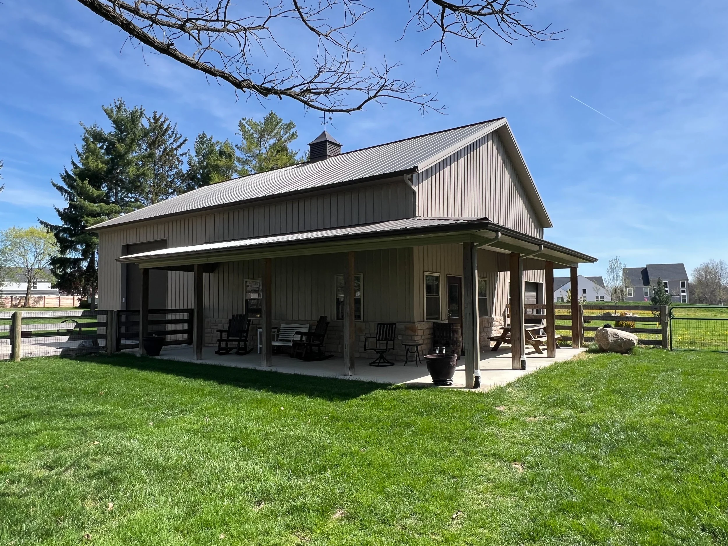 The image shows a house with a metal roof, a porch with chairs, potted plants, and surrounding green lawn. There are trees in the background and a partly cloudy sky with a visible contrail.