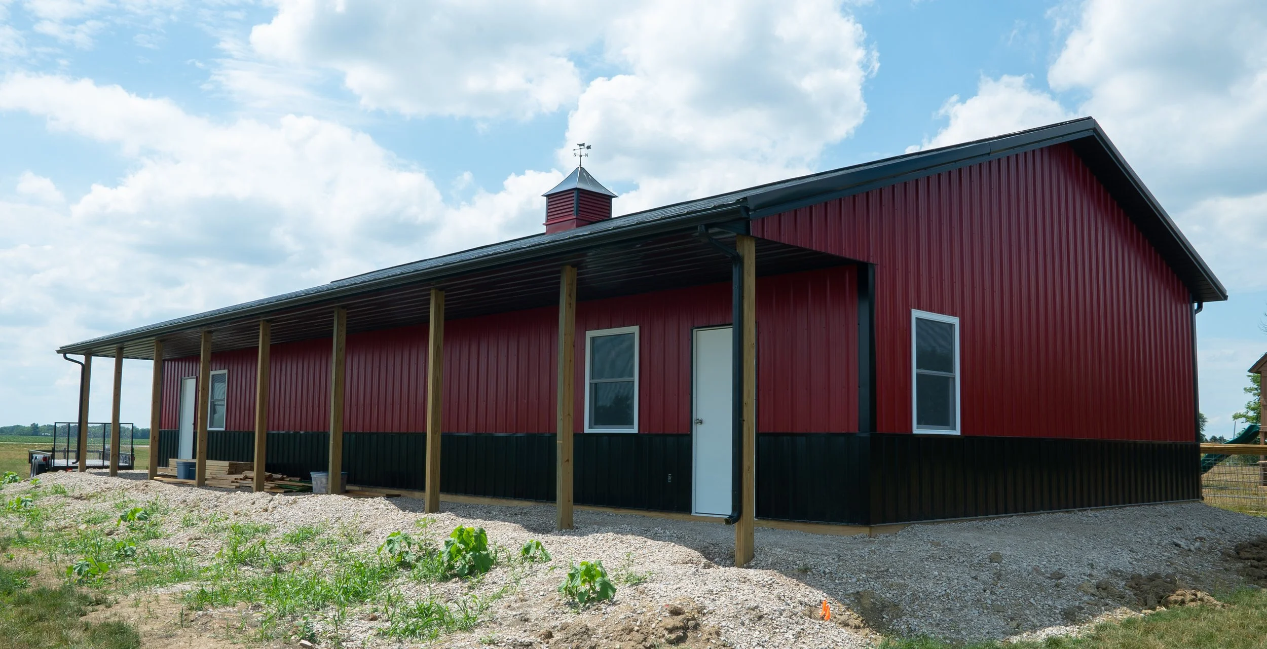 A red metal barn with a black lower section and white doors and windows, located in a rural area under a cloudy sky. The barn has a small cupola with a weather vane on top and a covered porch area supported by wooden posts.