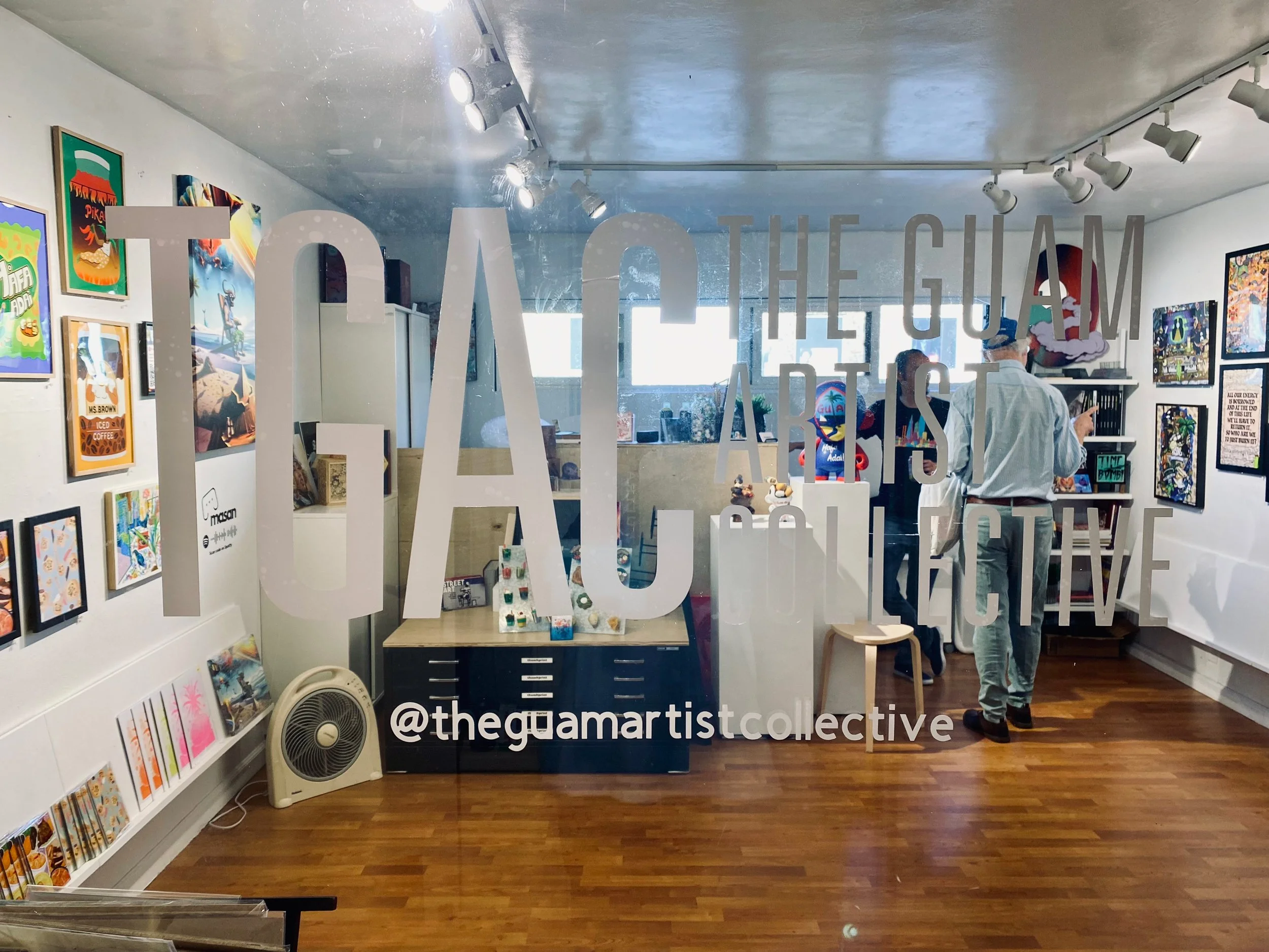 Inside an art gallery with various colorful paintings on the walls. Two people are looking at the artwork, one pointing at something. A fan is on the floor, and the gallery has a wooden floor. Text on the glass reads '@thegamaartiscollective'.