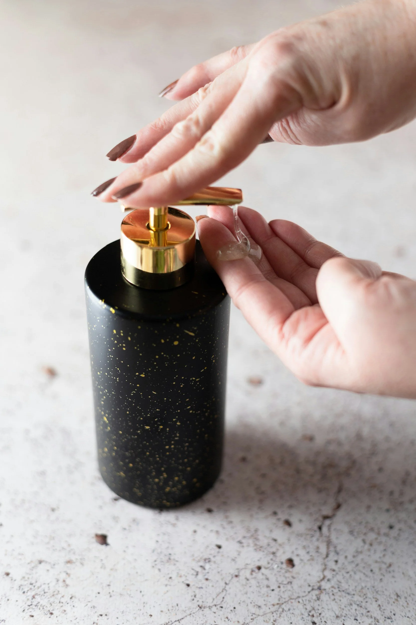 Person pressing a pump dispenser on a black bottle with gold speckles, dispensing a clear liquid onto their hand.