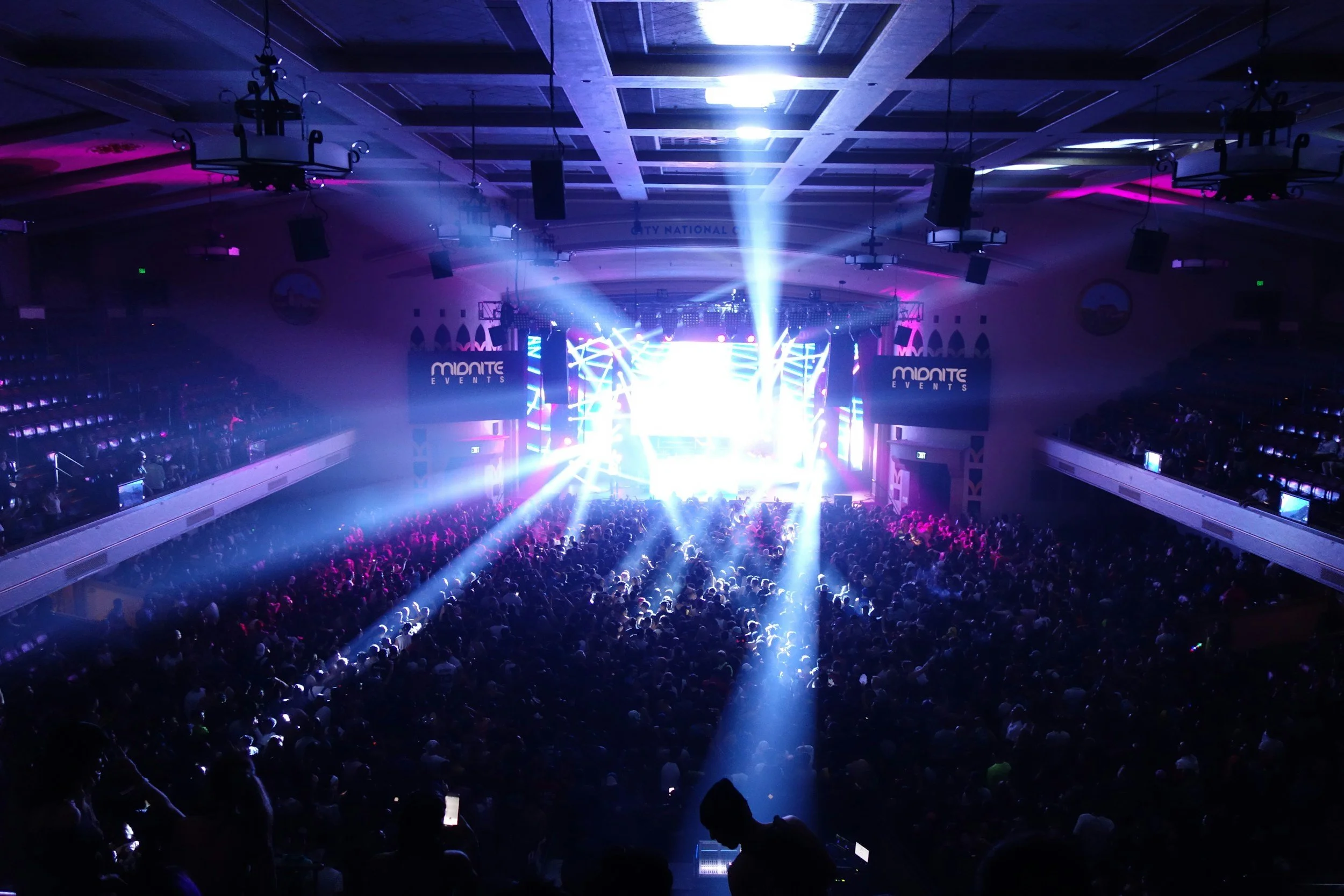 A large indoor concert or event venue filled with a crowded audience, colorful stage lighting, and large screens displaying 'Minitic Events'.