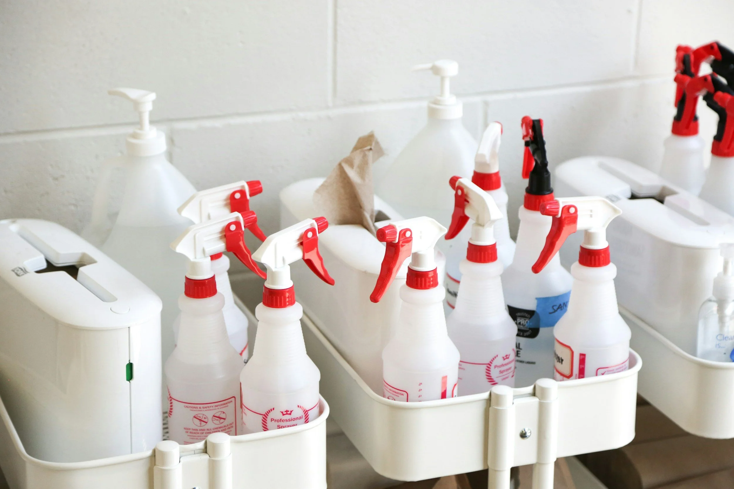 A white plastic cart with multiple spray bottles containing cleaning solutions, some with red and black spray nozzles, against a white tiled wall.