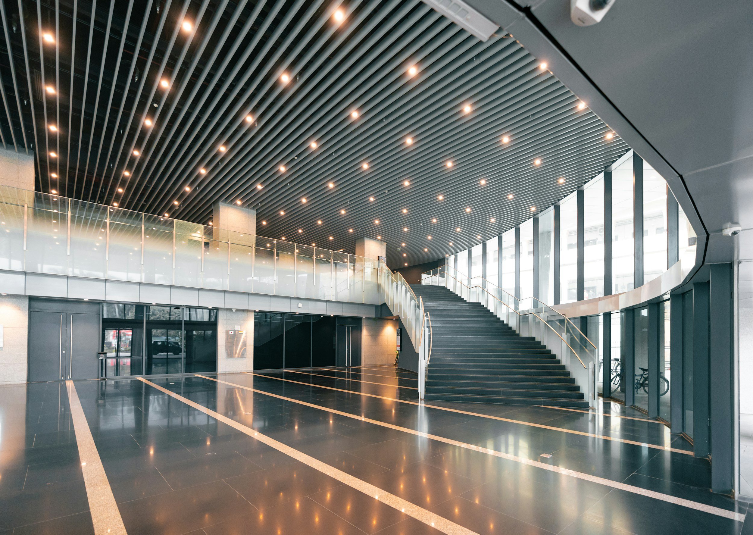 Modern indoor lobby with a staircase, black tiled floor, large windows, metal railing, and a ceiling with evenly spaced small lights.