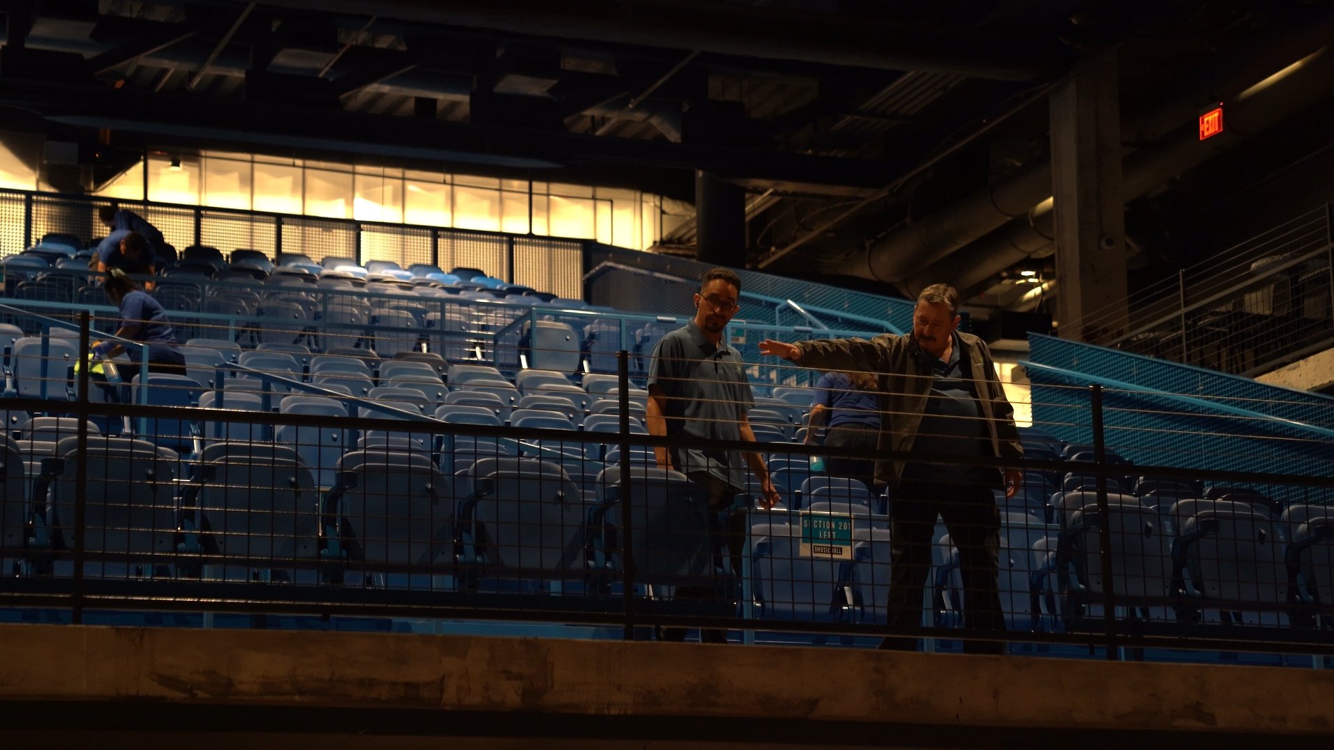 Two men in casual clothing standing and talking on the upper level of a stadium with blue seats, while staff members are setting up in the background.