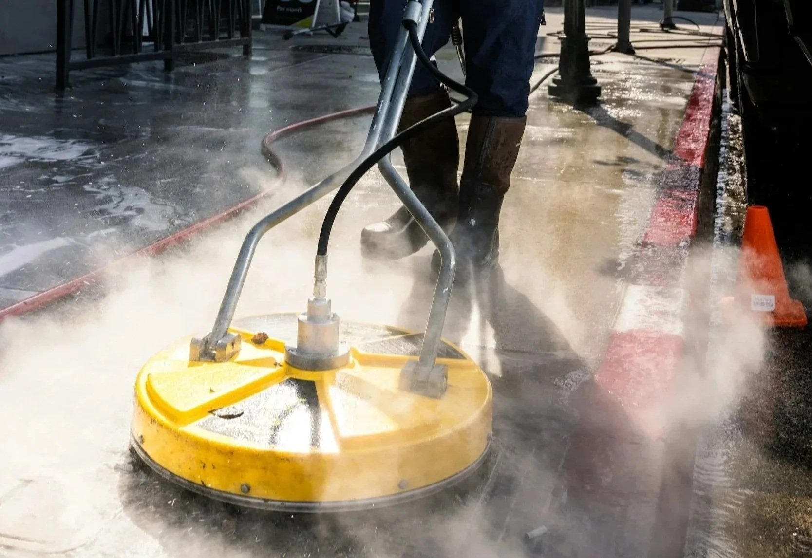 Person operating a floor cleaning machine on a wet sidewalk at night, with chairs and a table in the background and orange traffic cones nearby.