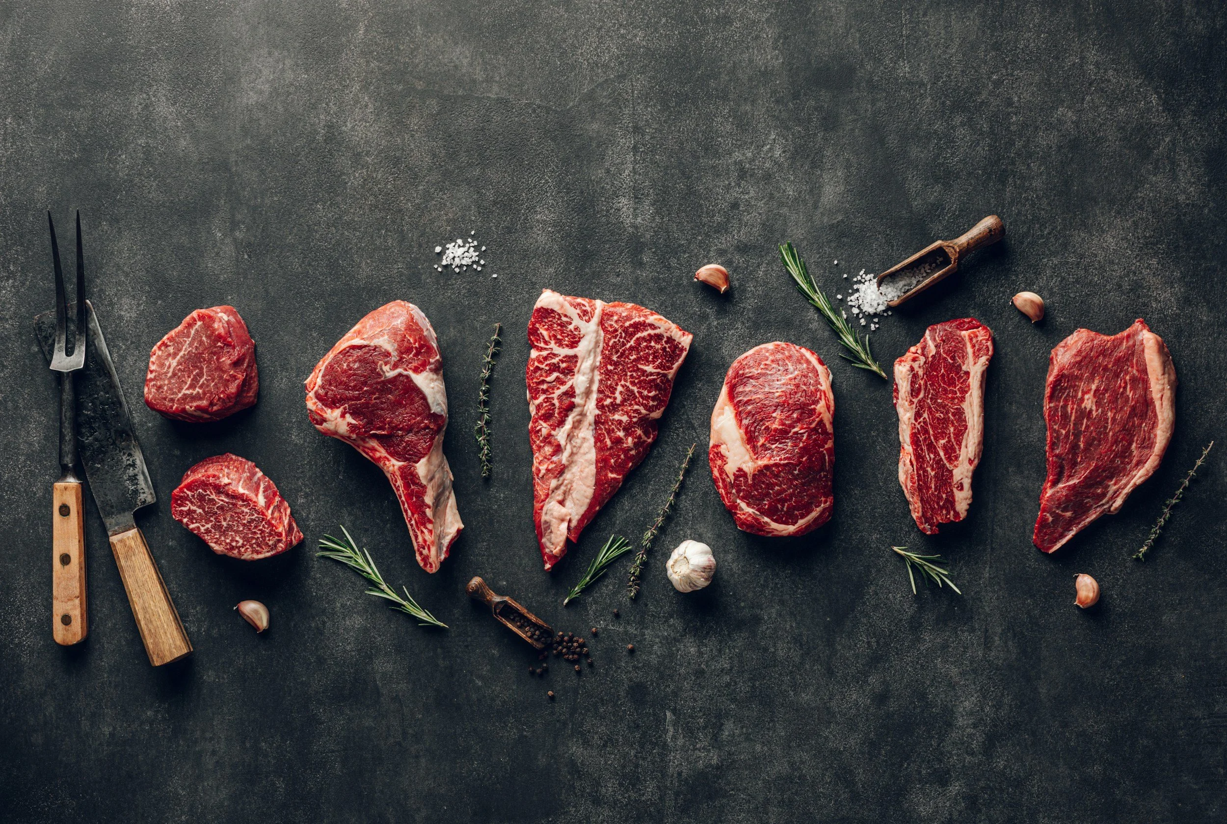 Assorted raw beef steaks arranged on a dark textured surface, accompanied by herbs, garlic, salt, black pepper, and barbecuing tools.