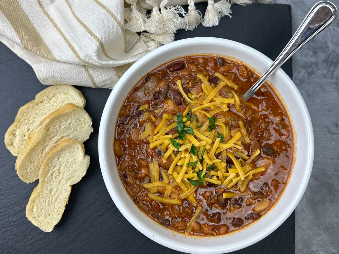A bowl of chili with shredded cheese and chopped parsley, served with sliced bread on a black surface.