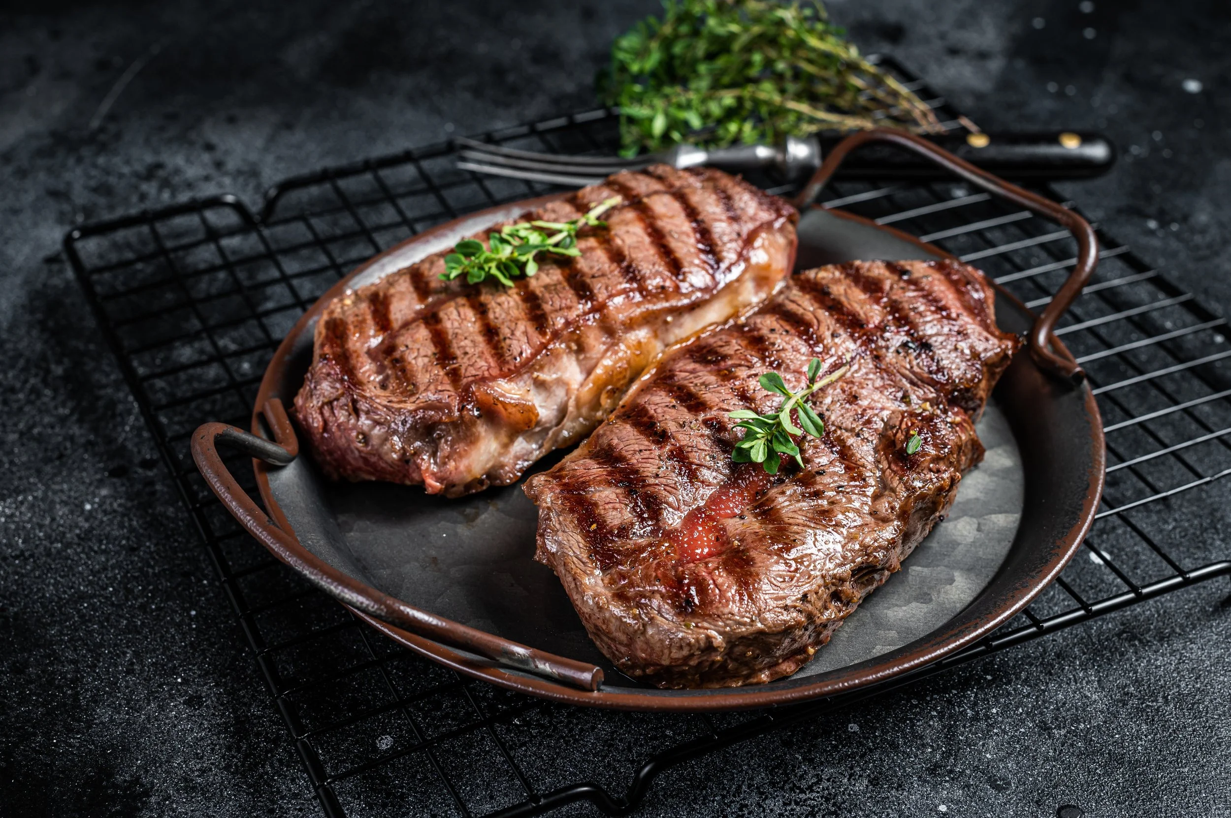 Two grilled steaks garnished with fresh herbs on a round cast iron skillet, placed on a dark textured surface with a cooling rack underneath.
