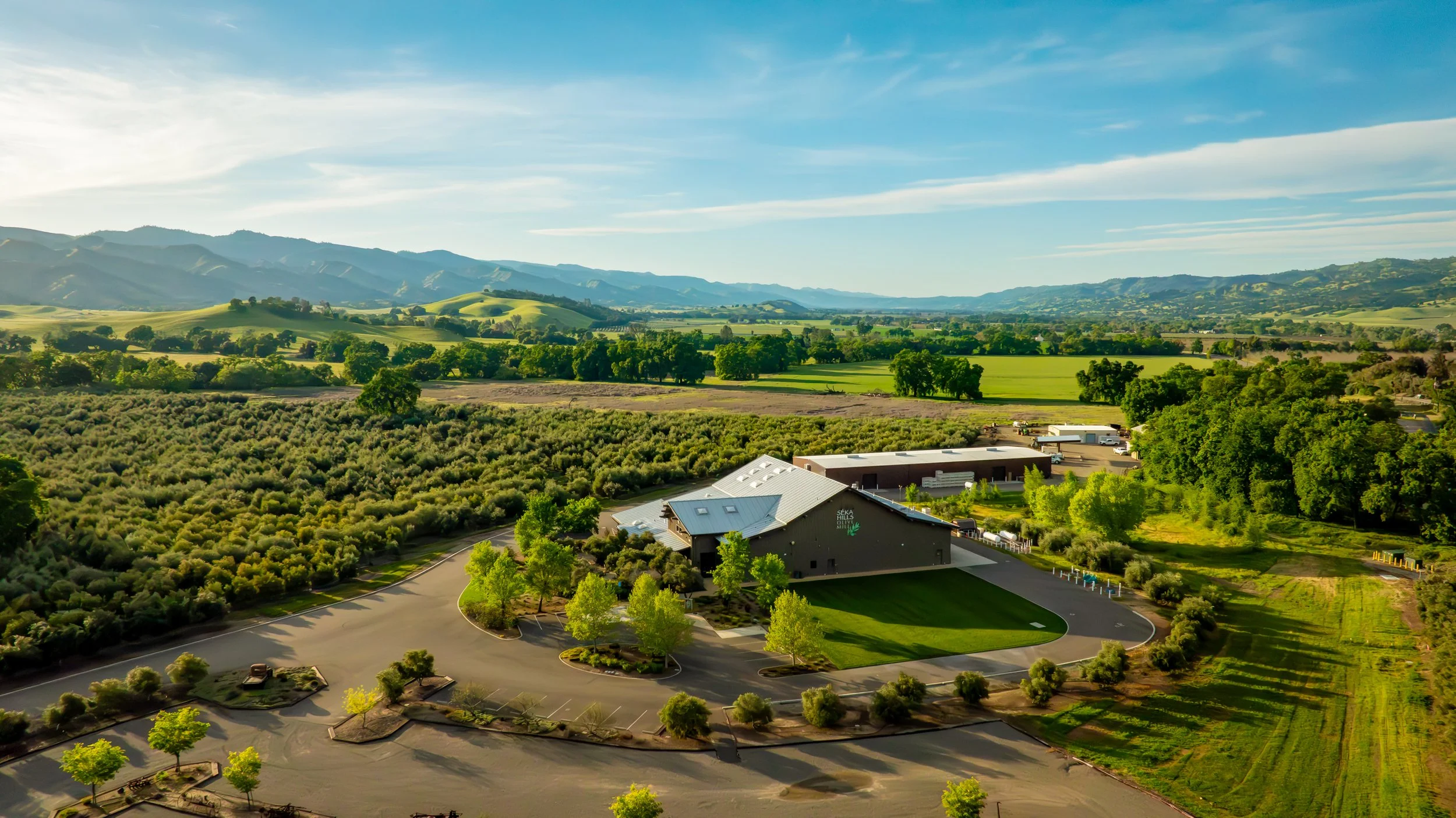 A scenic view of rolling green hills, trees, a parking lot, and buildings under a partly cloudy sky.