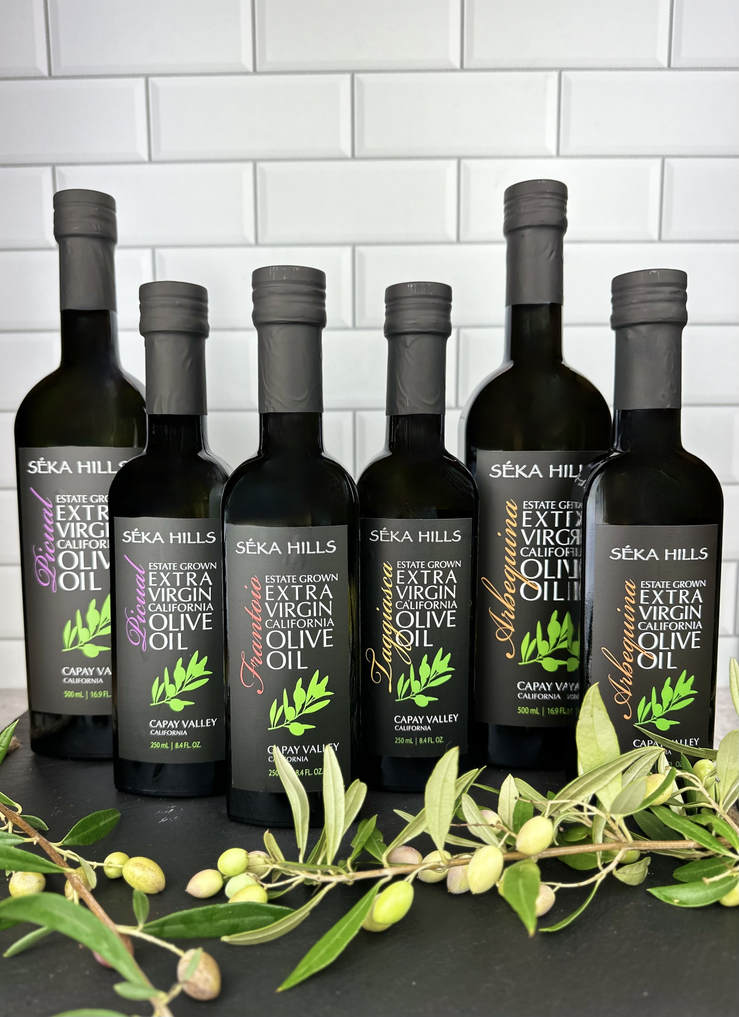 Six bottles of Séka Hills extra virgin olive oil arranged on a black surface with green olive branches in the foreground and a white tiled wall in the background.
