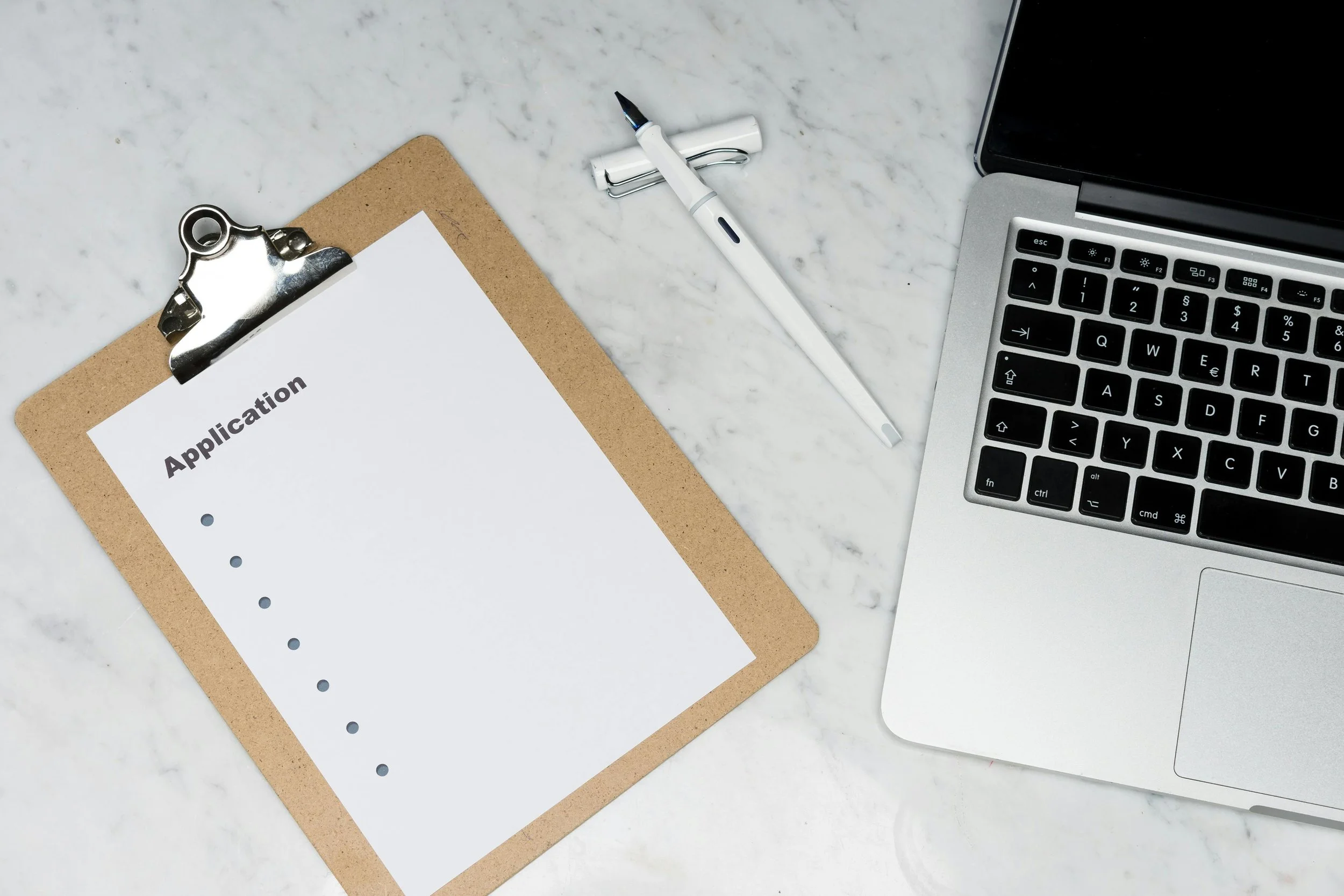 A clipboard reading "Application" sitting next to an ink pen and a partially visible MacBook