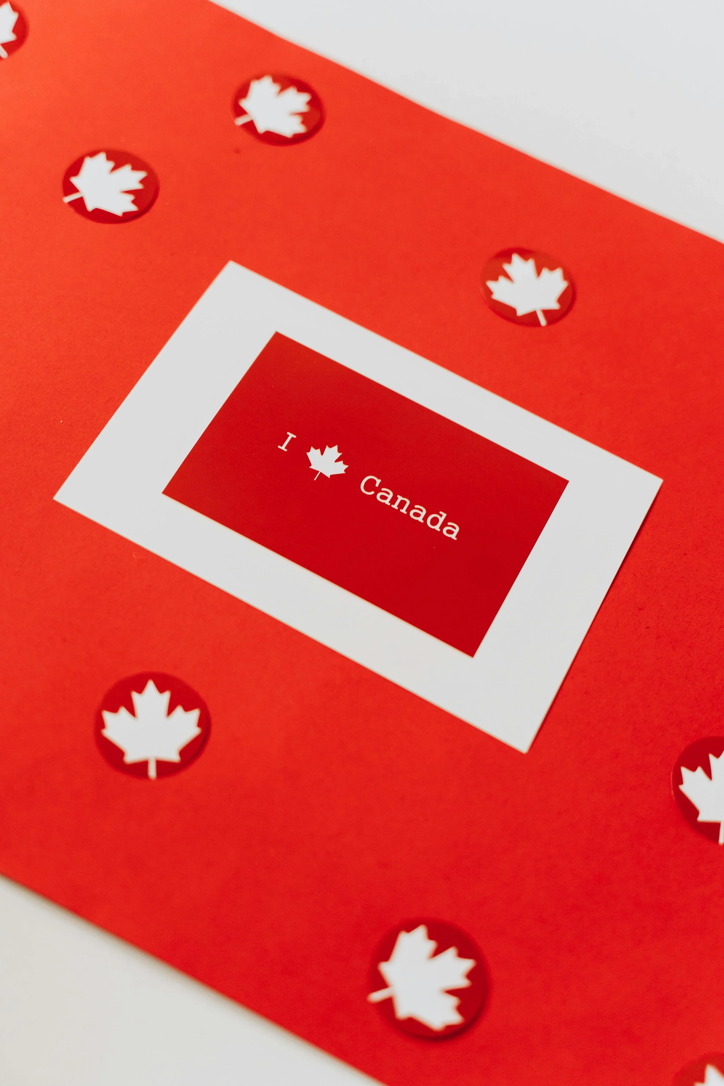 a clover-shaped picture of a red background with white maple leaves. In the center, there is a red card with a wide white border that says “I” maple leaf “Canada” in white letters
