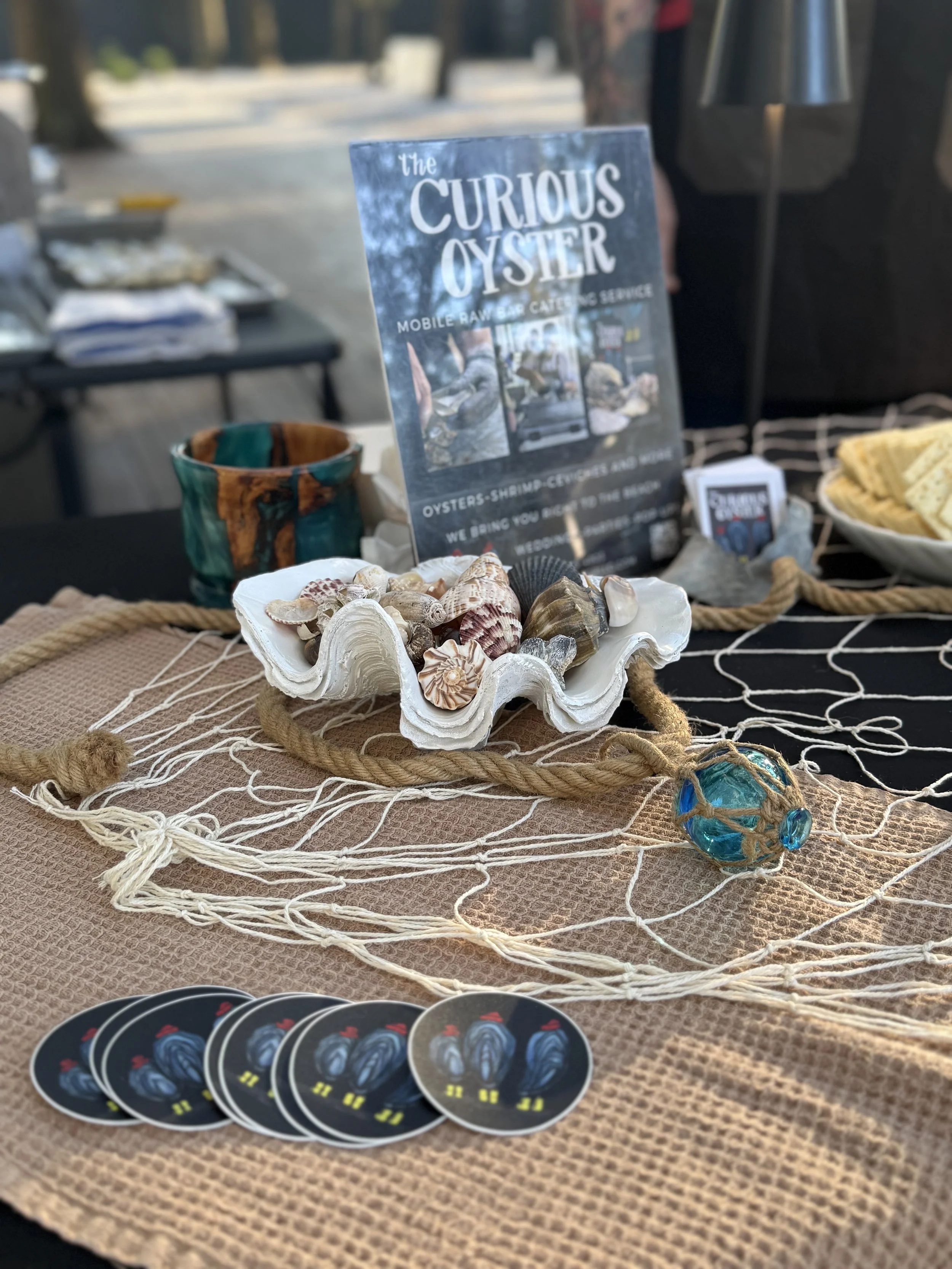 Display of seashells on a white shell-shaped dish, a set of circular black tokens with blue and yellow design, a sign for 'the curious oyster' with text and images, a colorful small glass ball, and various other items on a textured tan and black surface.