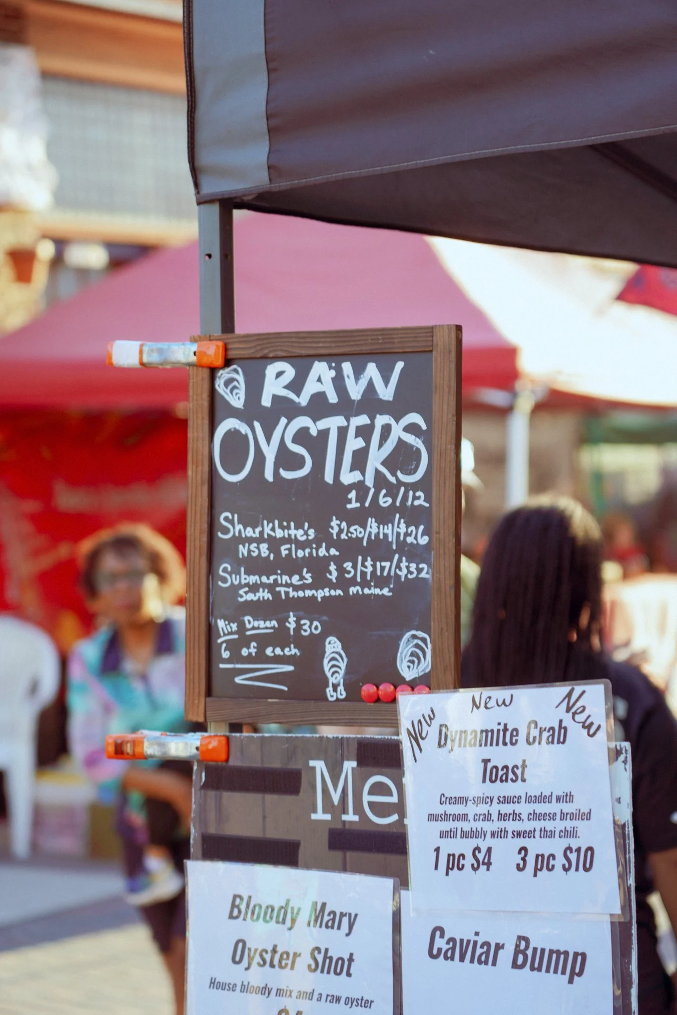 A signboard listing raw oysters and other seafood items at an outdoor market, with other market booths and blurred people in the background.