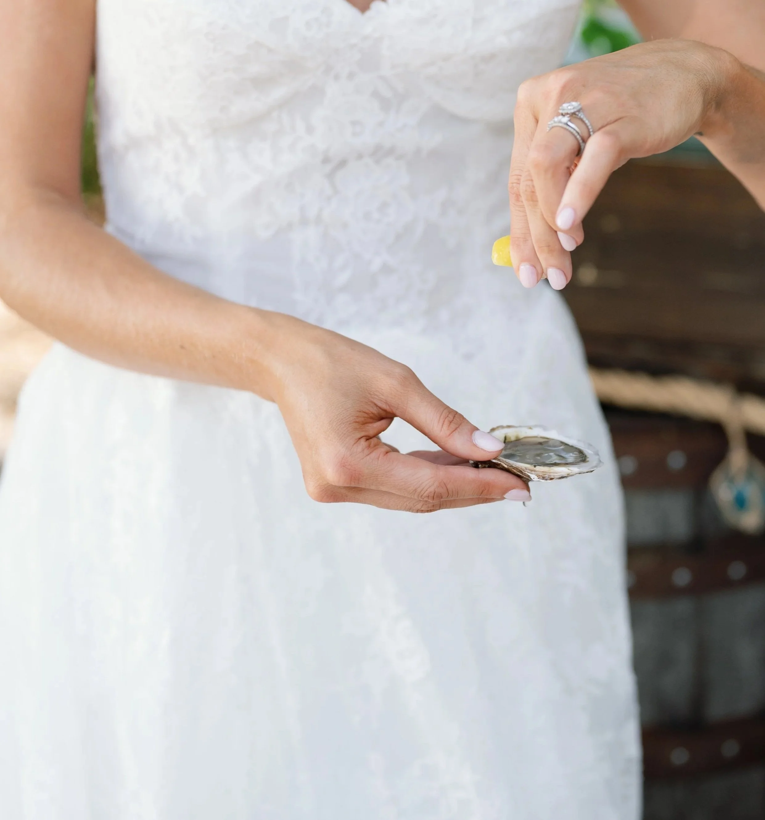 Close-up of a woman’s hands, holding a large oyster shell with an oyster inside, and touching it with her finger. She is wearing a white dress and a diamond ring on her right hand.