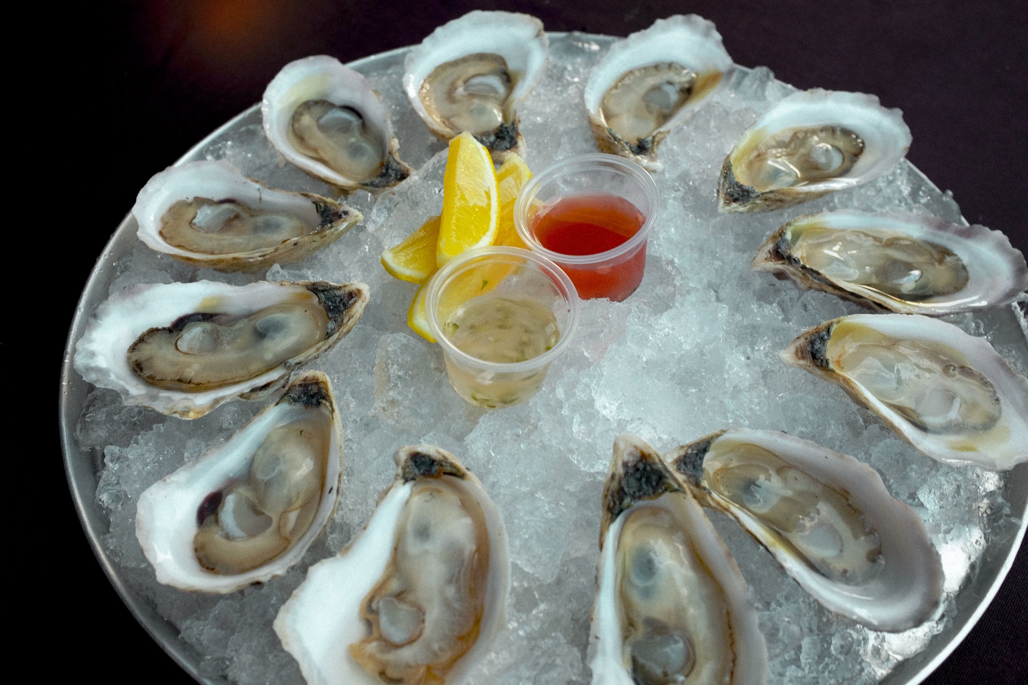 A platter of fresh oysters on ice with lemon wedges, cocktail sauce, and mignonette sauce.