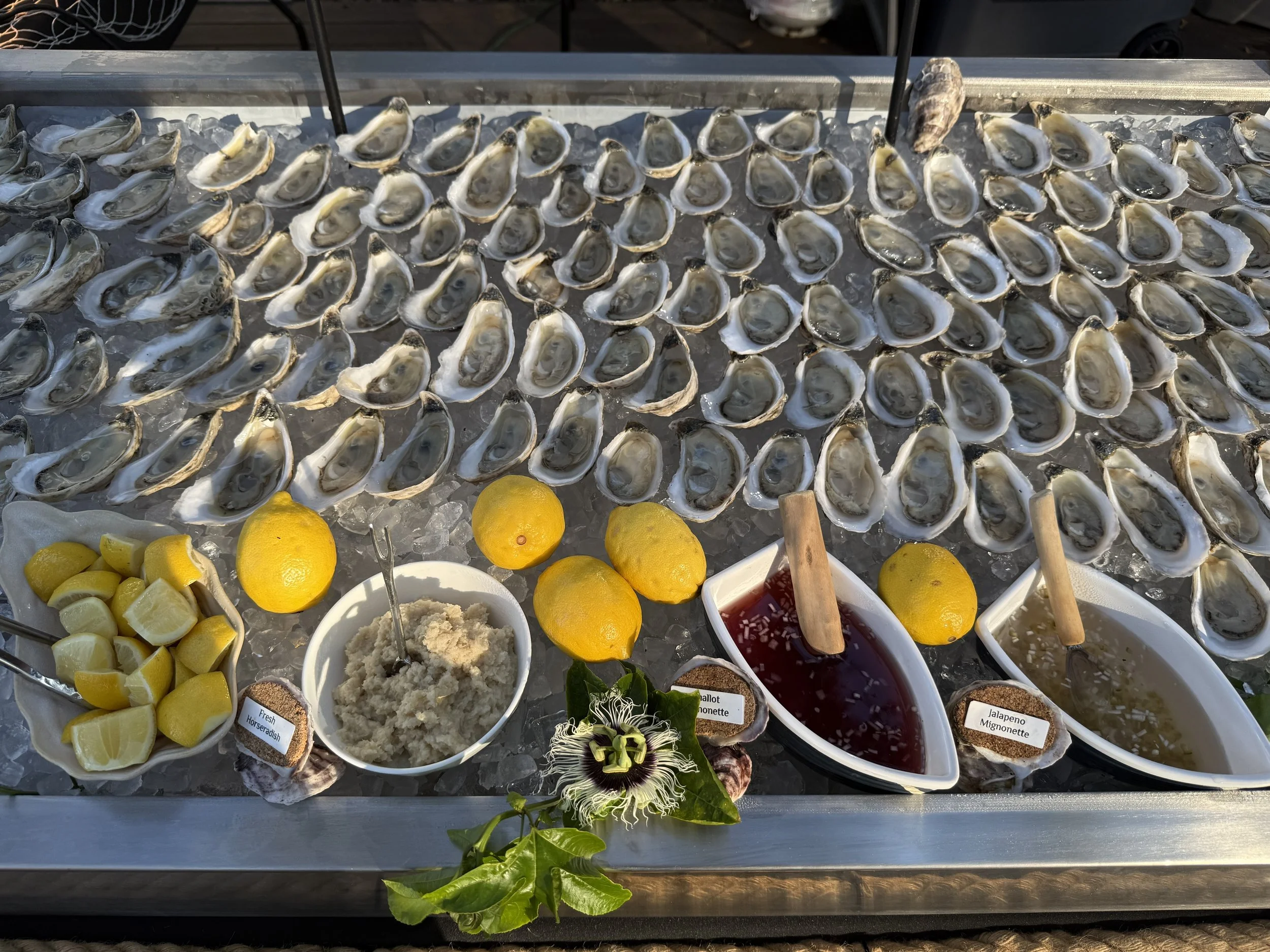 A display of oysters on ice with lemon wedges and various dipping sauces, set on a metal table at an outdoor seafood market.