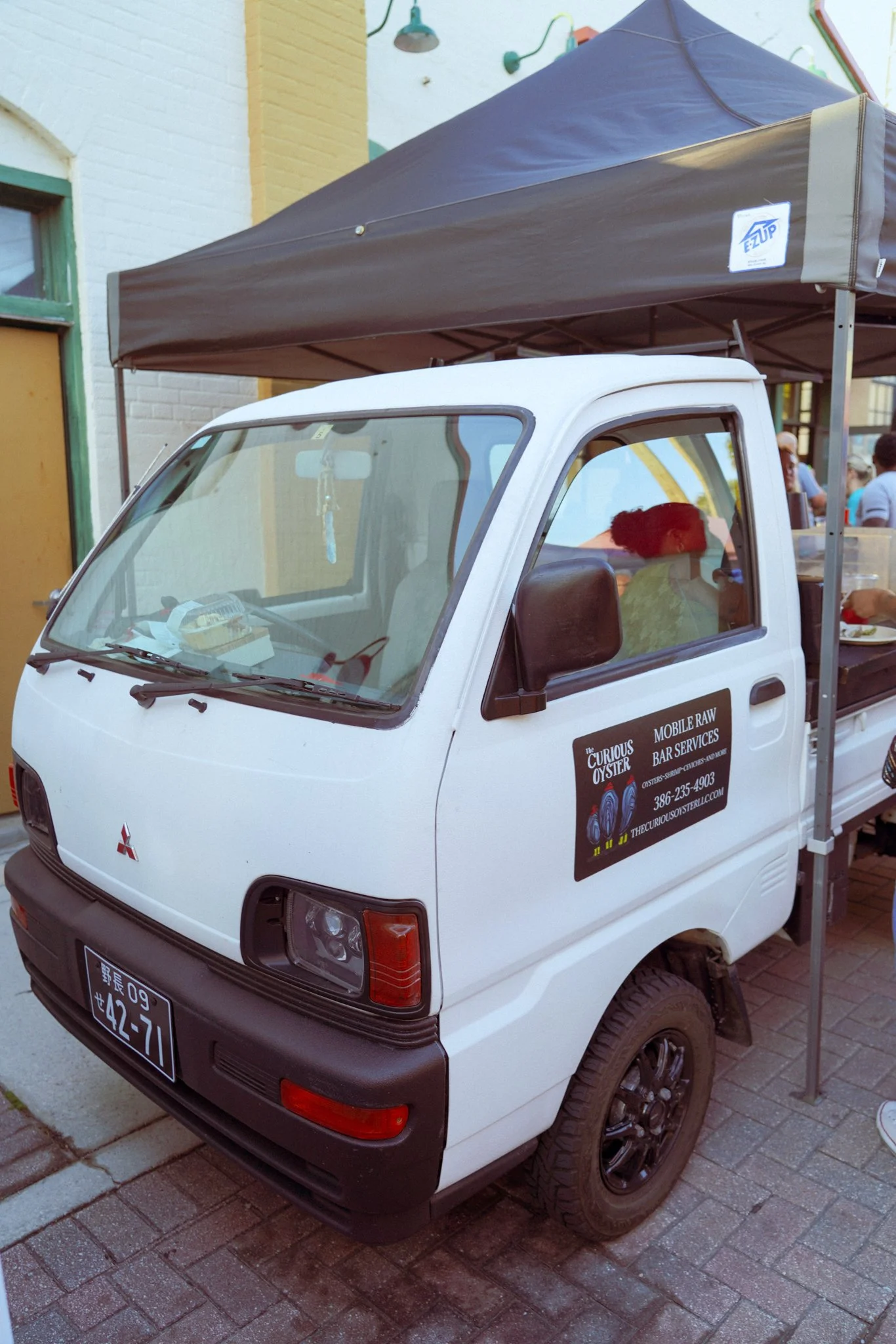 A small white Mitsubishi mini truck with a black bumper and black wheels parked on a brick pavement. The truck has a sign on the door that reads 'Curious Oyster' with additional text about mobile raw bar services and a phone number. The truck has a canopy attached to the back.