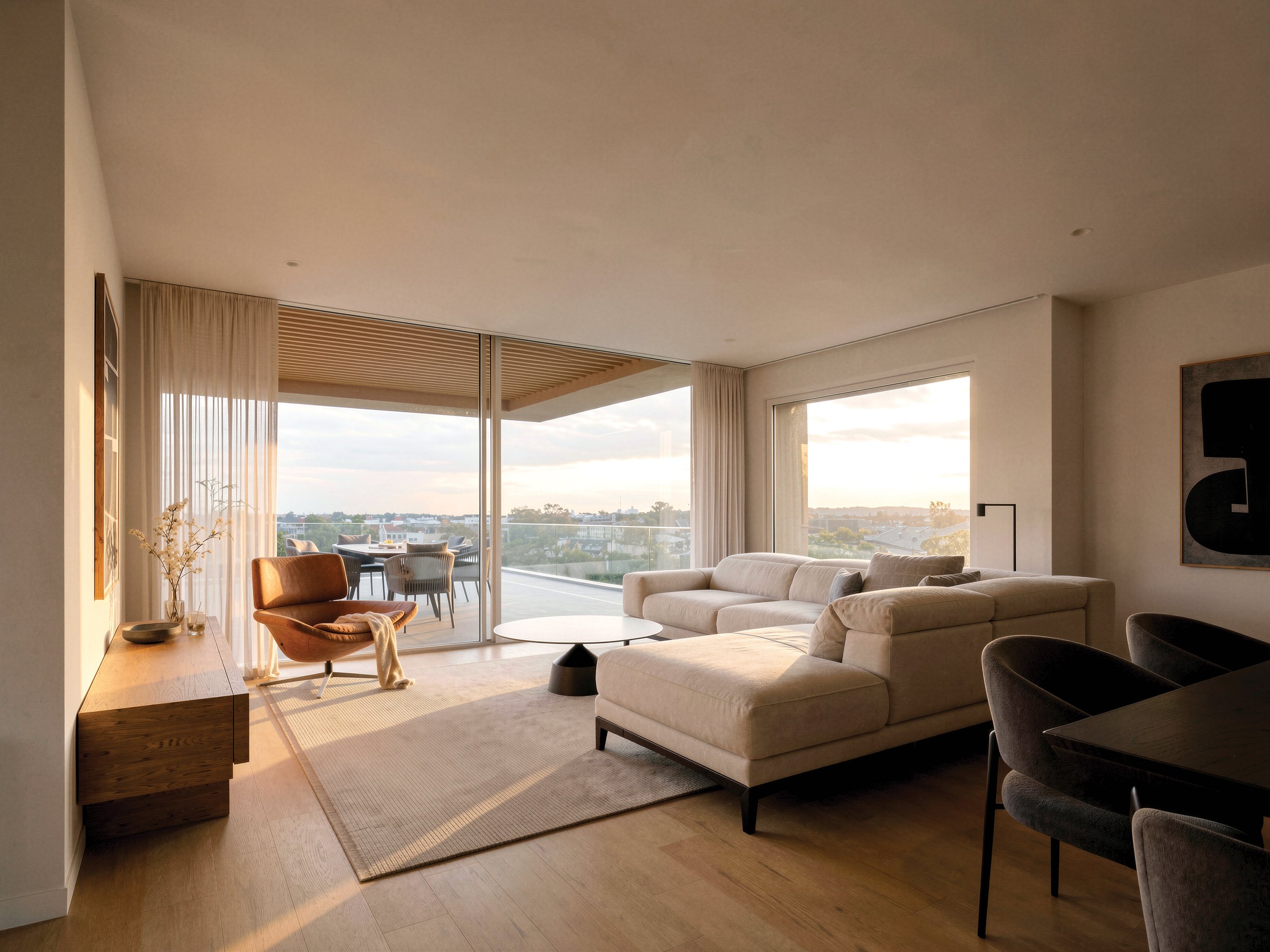 Living room with large glass sliding door opening to balcony, view of cityscape and sky, beige sofa, armchair, coffee table, wooden sideboard, curtains, artwork, and modern decor.
