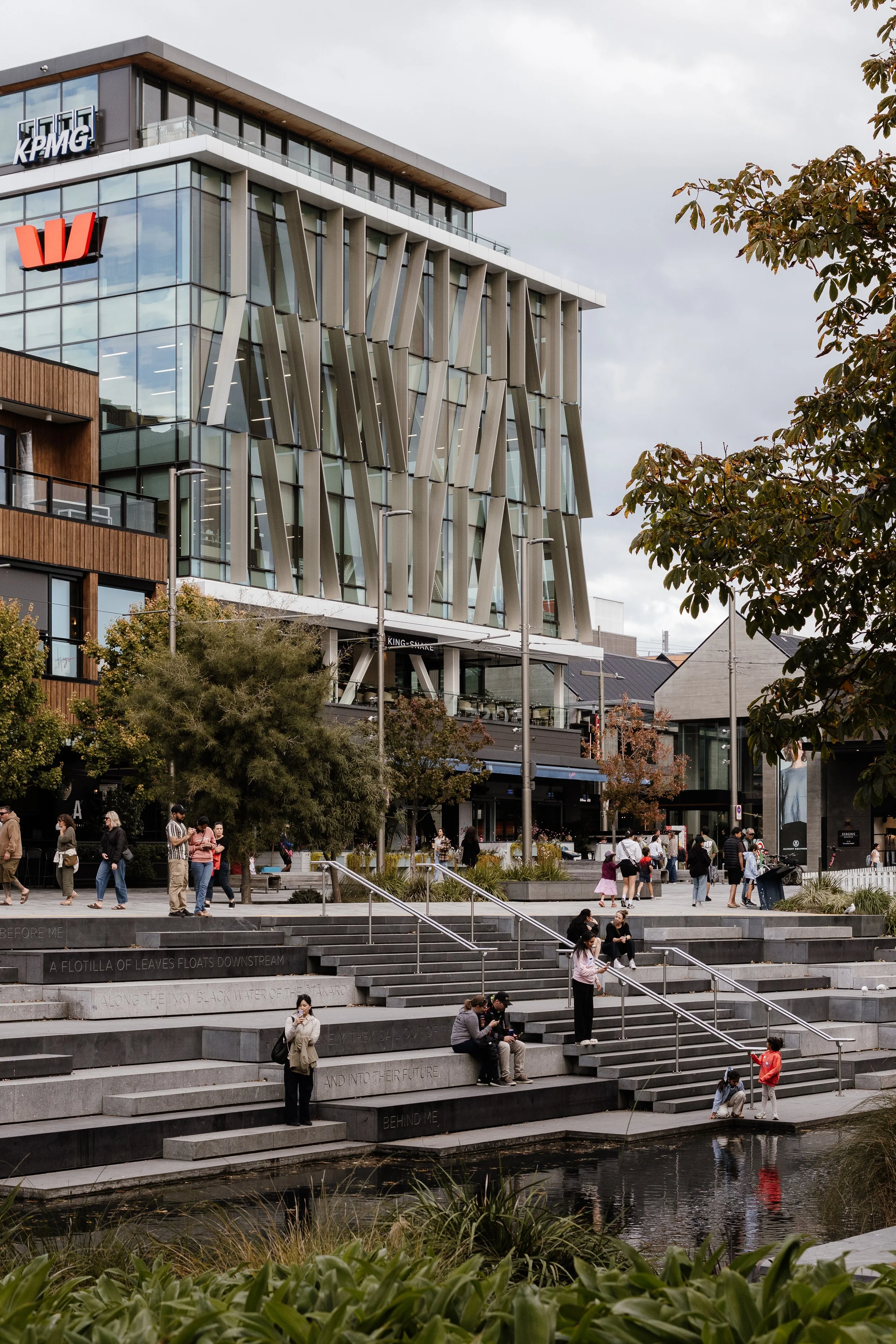 People gather on wide outdoor steps near a water feature in an urban area, with modern glass and metal buildings, including a KPMG office and a Westpac bank branch, in the background. Some people are sitting, walking, and talking, with trees and lamp posts around the area.
