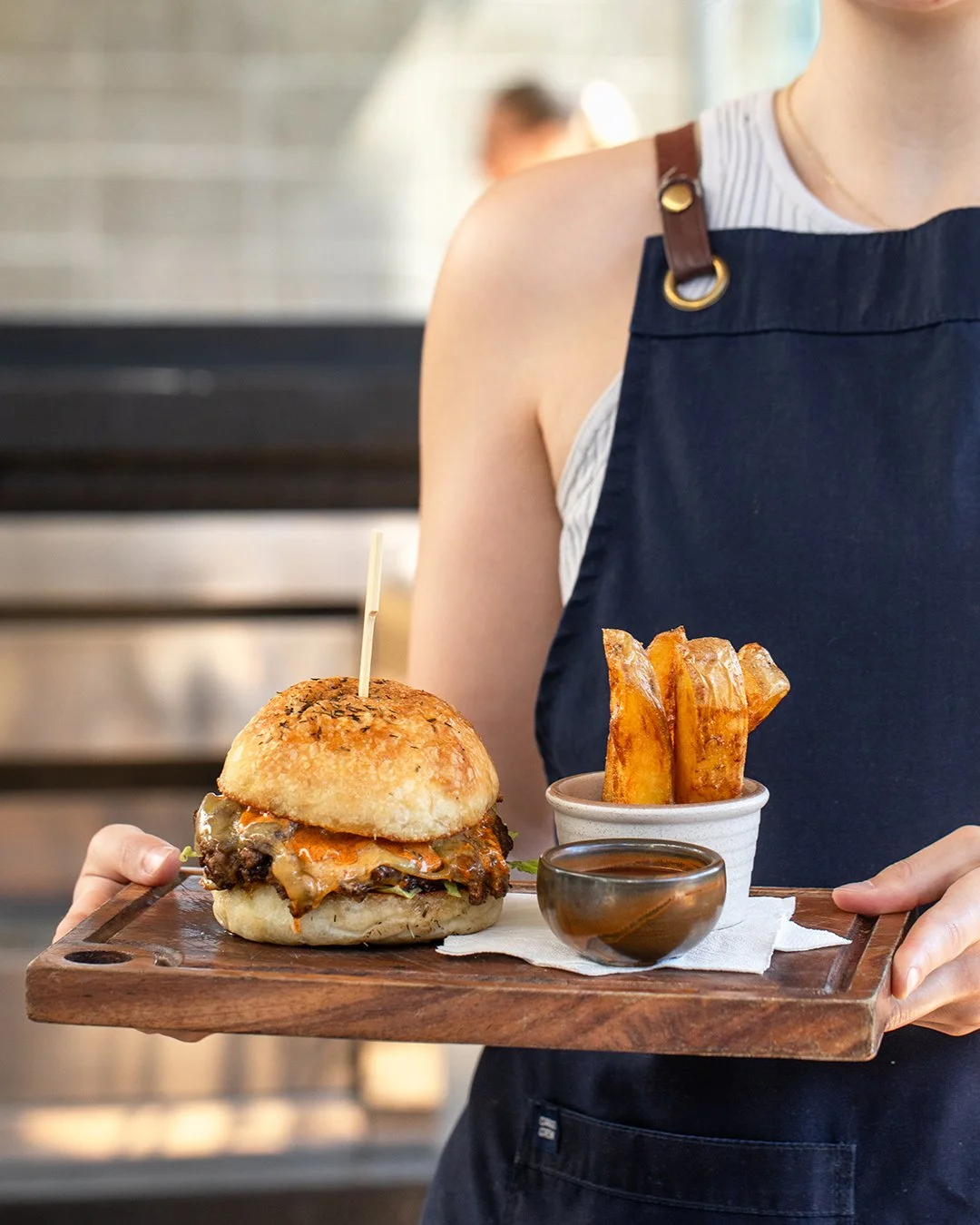 Person holding a wooden tray with a burger, sweet potato fries, and a small bowl of dipping sauce, in a restaurant setting.