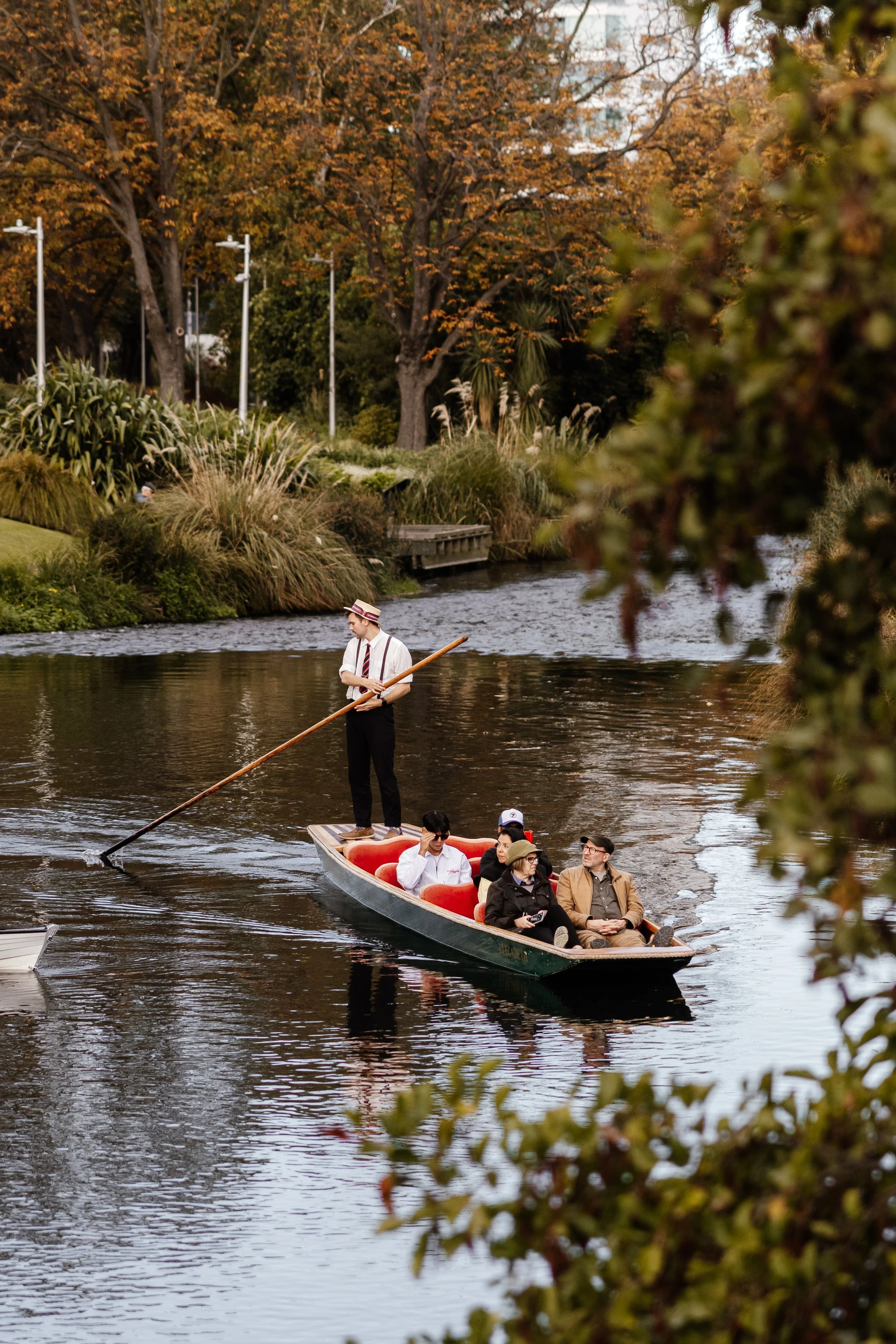 People enjoying a boat ride on a river surrounded by trees with autumn foliage, some with orange leaves, and a person standing on a paddleboard steering the boat.