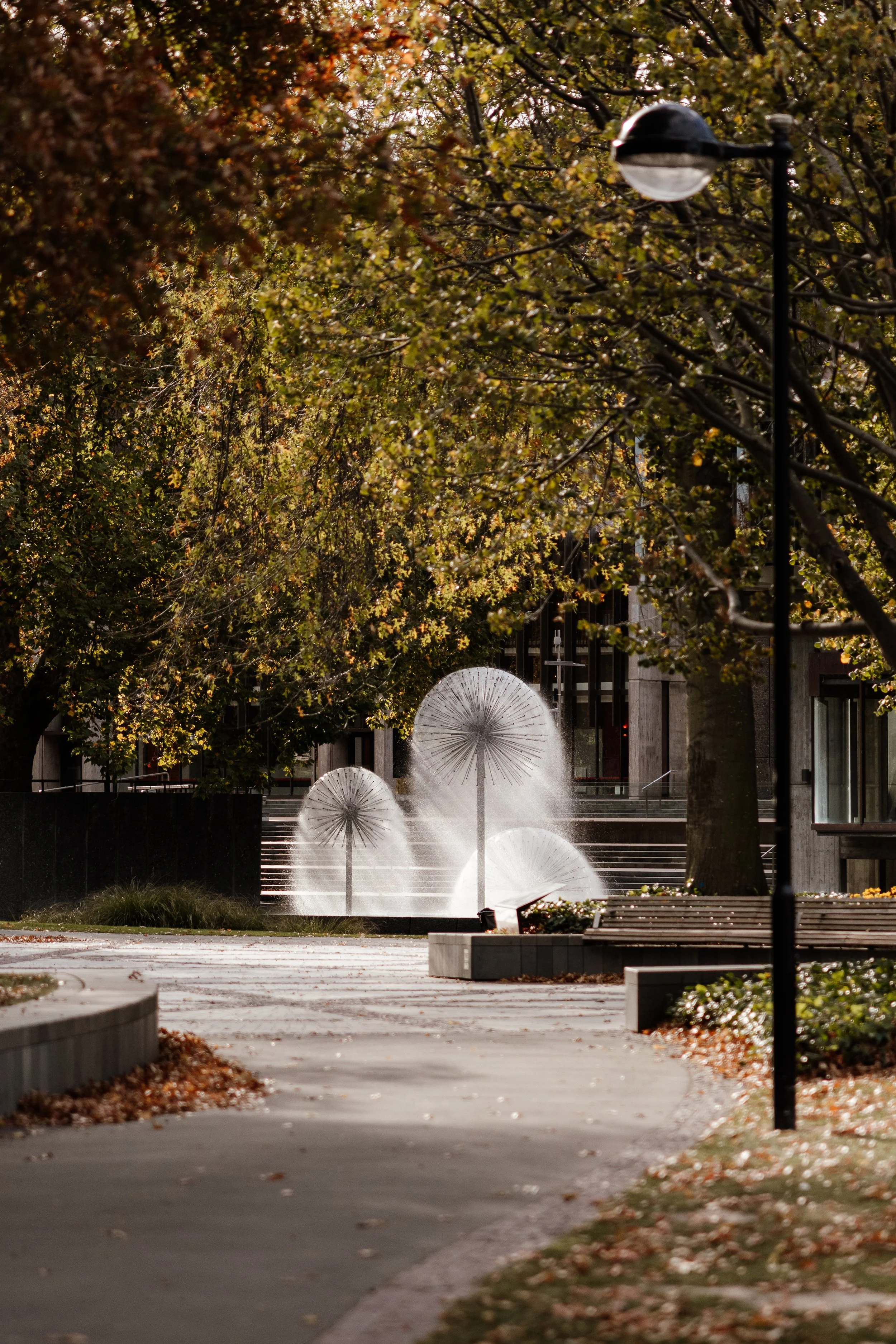 A park scene at dusk with trees showing autumn leaves, a modern street lamp, benches, and four umbrella-shaped water fountains shooting water upward.
