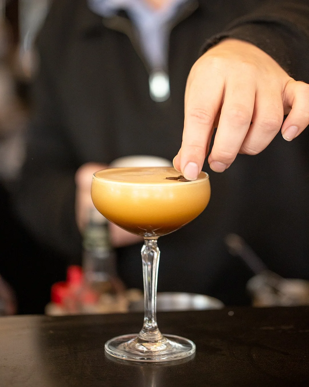 Close-up of a hand decorating a cocktail with a coffee bean in a coupe glass filled with a layered brown and creamy drink.