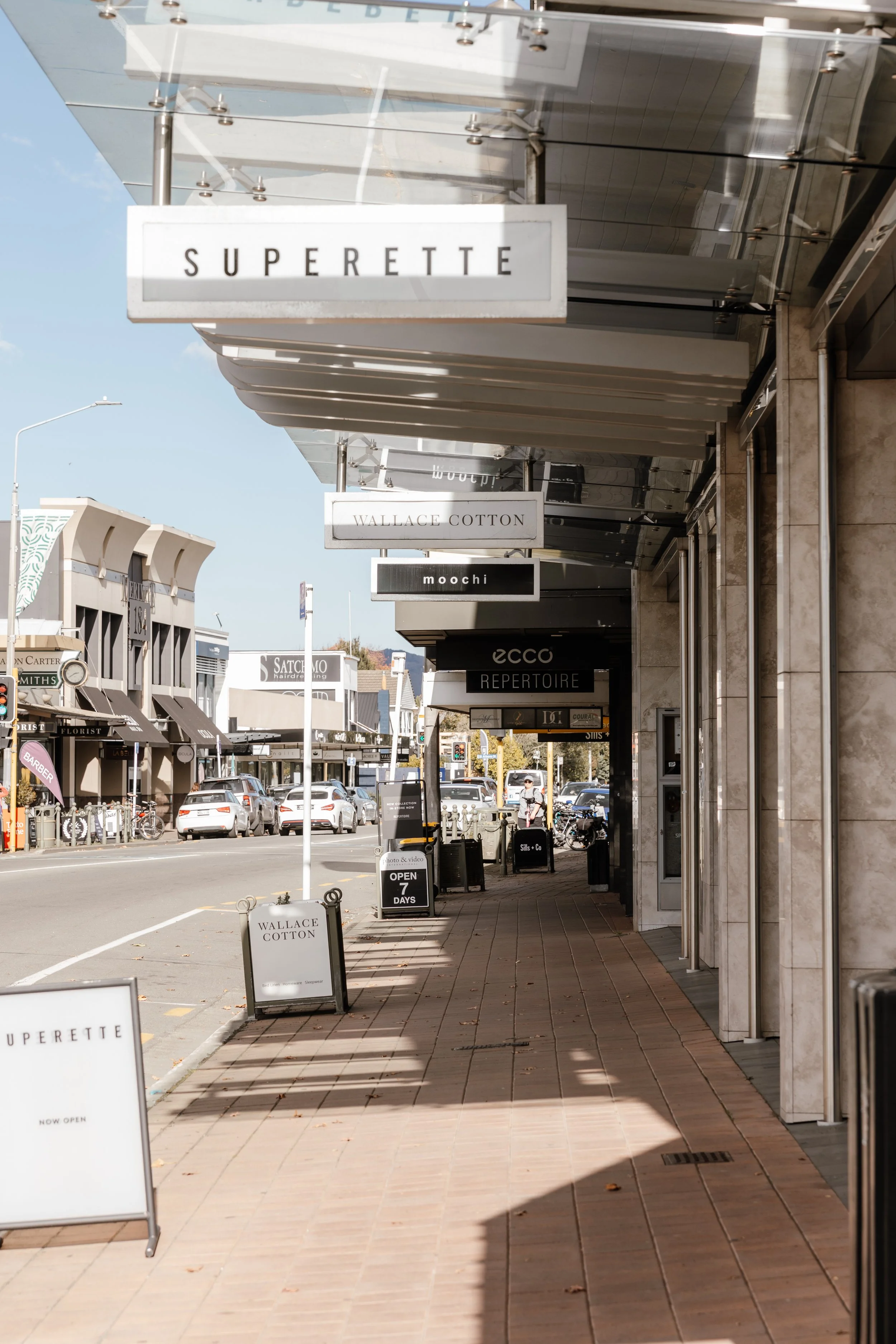 A sidewalk outside retail stores with signs for Wallace Cotton, Superette, Moochi, and ECCO, along with sidewalk signs indicating stores are open 7 days.