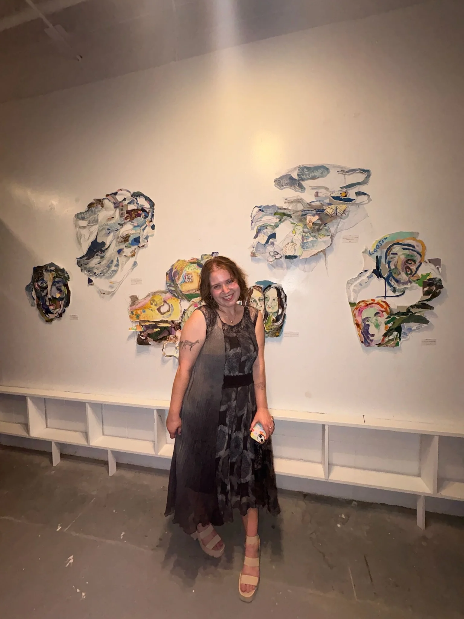 A woman standing in front of a wall with colorful abstract artwork, smiling, holding a phone, wearing a sleeveless dress and high-heeled sandals.