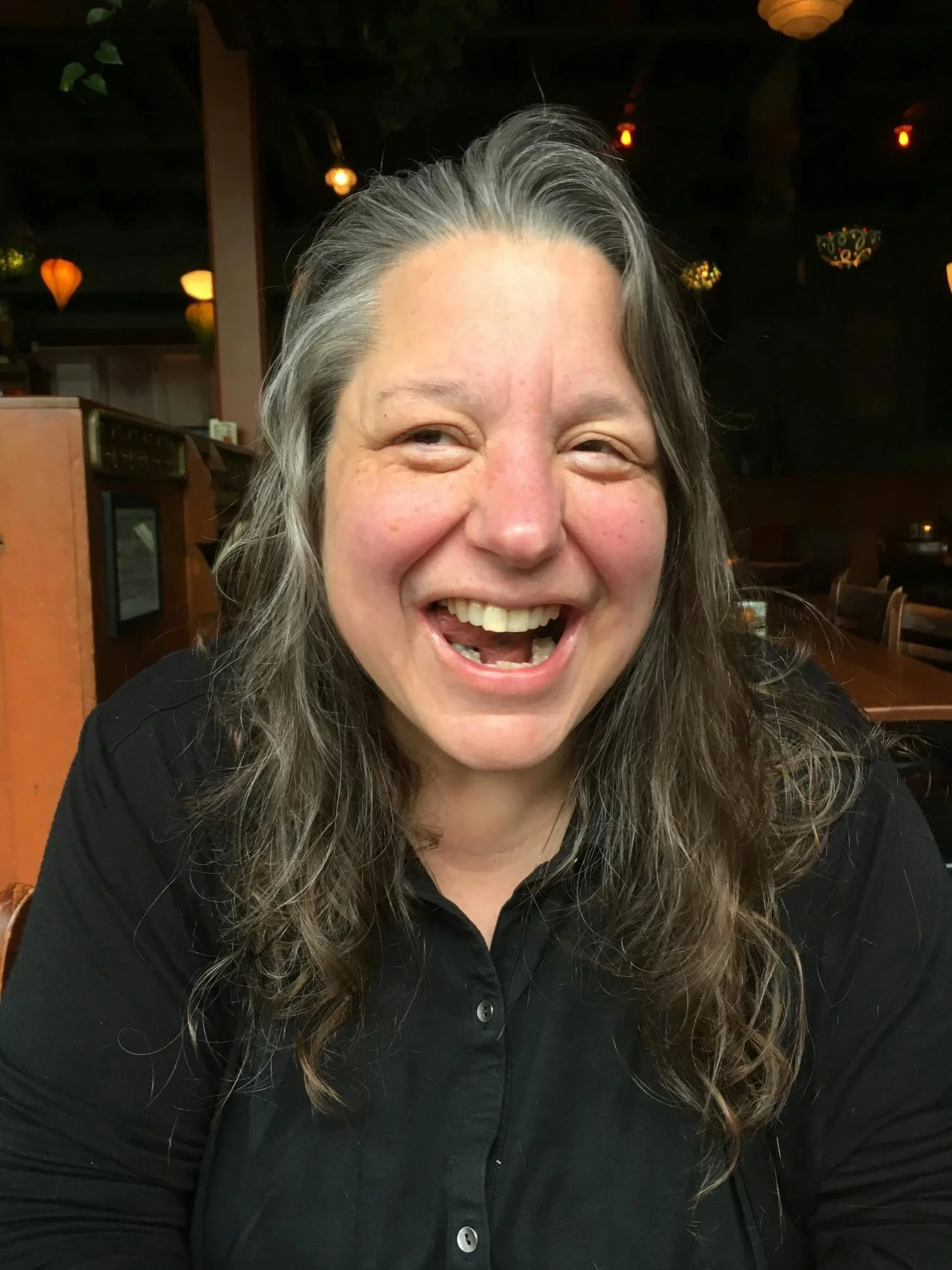 Close-up of a smiling woman with long gray hair, wearing a black button-up shirt, in a dimly lit restaurant with warm hanging lights in the background.