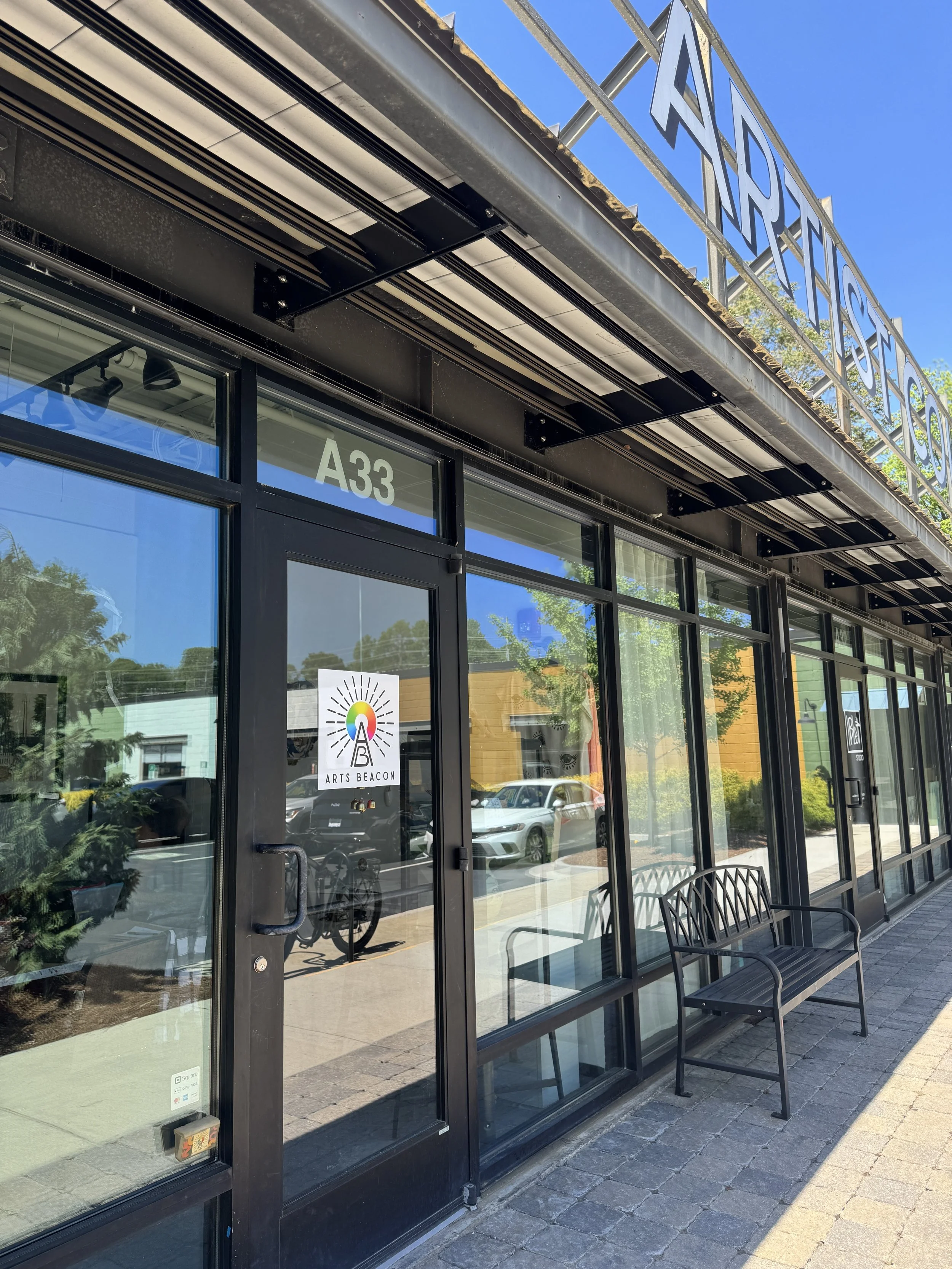 Exterior view of a storefront with large glass windows and a door, marked with the number A33, and a sign reading 'Arts Beacon' with a rainbow-colored beacon icon. Two black benches are outside. The area appears to be in a shopping district with other buildings and green trees reflected in the windows, under a blue sky.