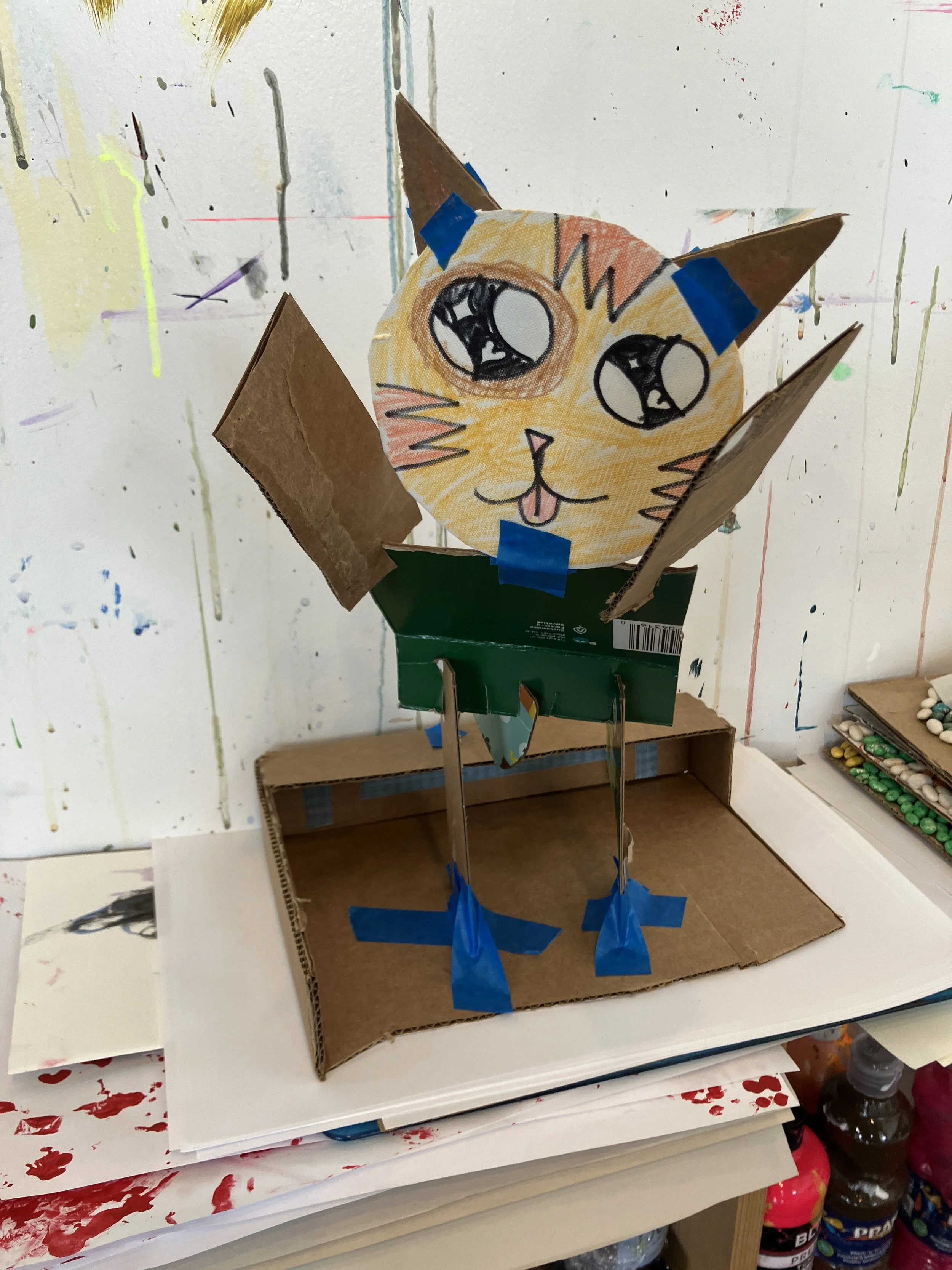 A handmade cardboard and paper craft of Pikachu from Pokémon with a drawn face on a paper circle, taped onto a cardboard body, and set on a white surface with art supplies and paint bottles around.