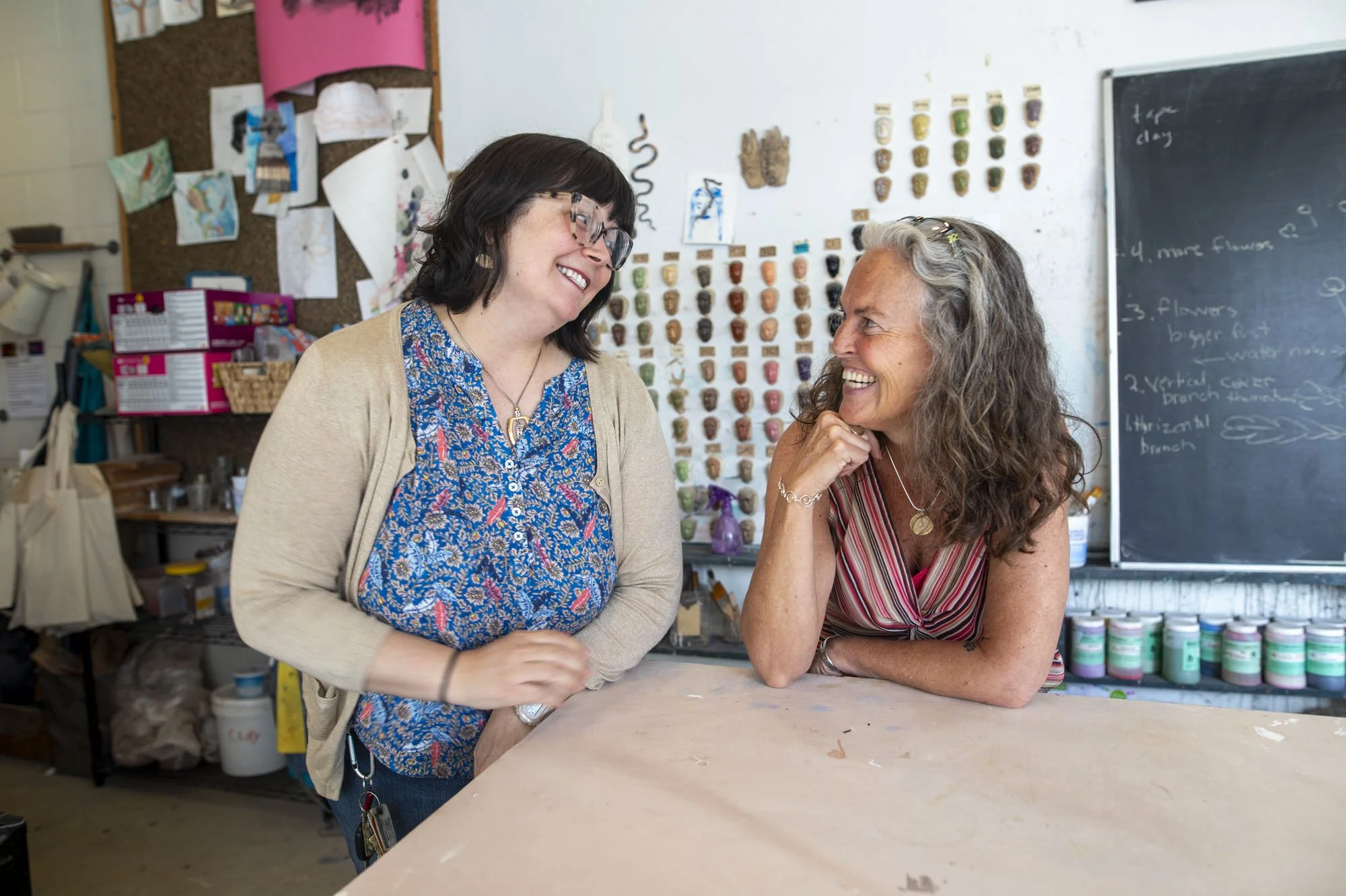 Two women are smiling and engaging in conversation in a classroom or studio with art supplies and decor in the background.