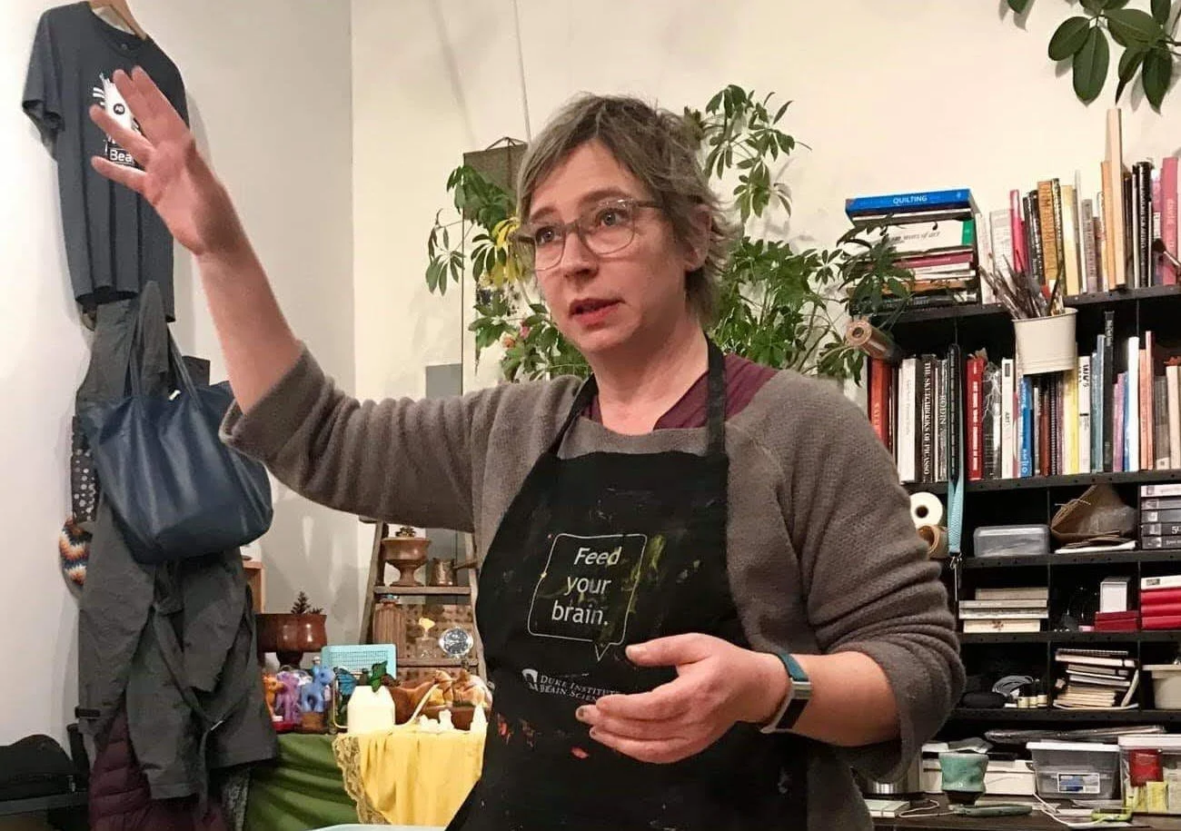 A woman with gray hair and glasses wearing a black apron with the words 'Feed your brain' stands in a room with bookshelves, plants, and various items, gesturing with her right hand.