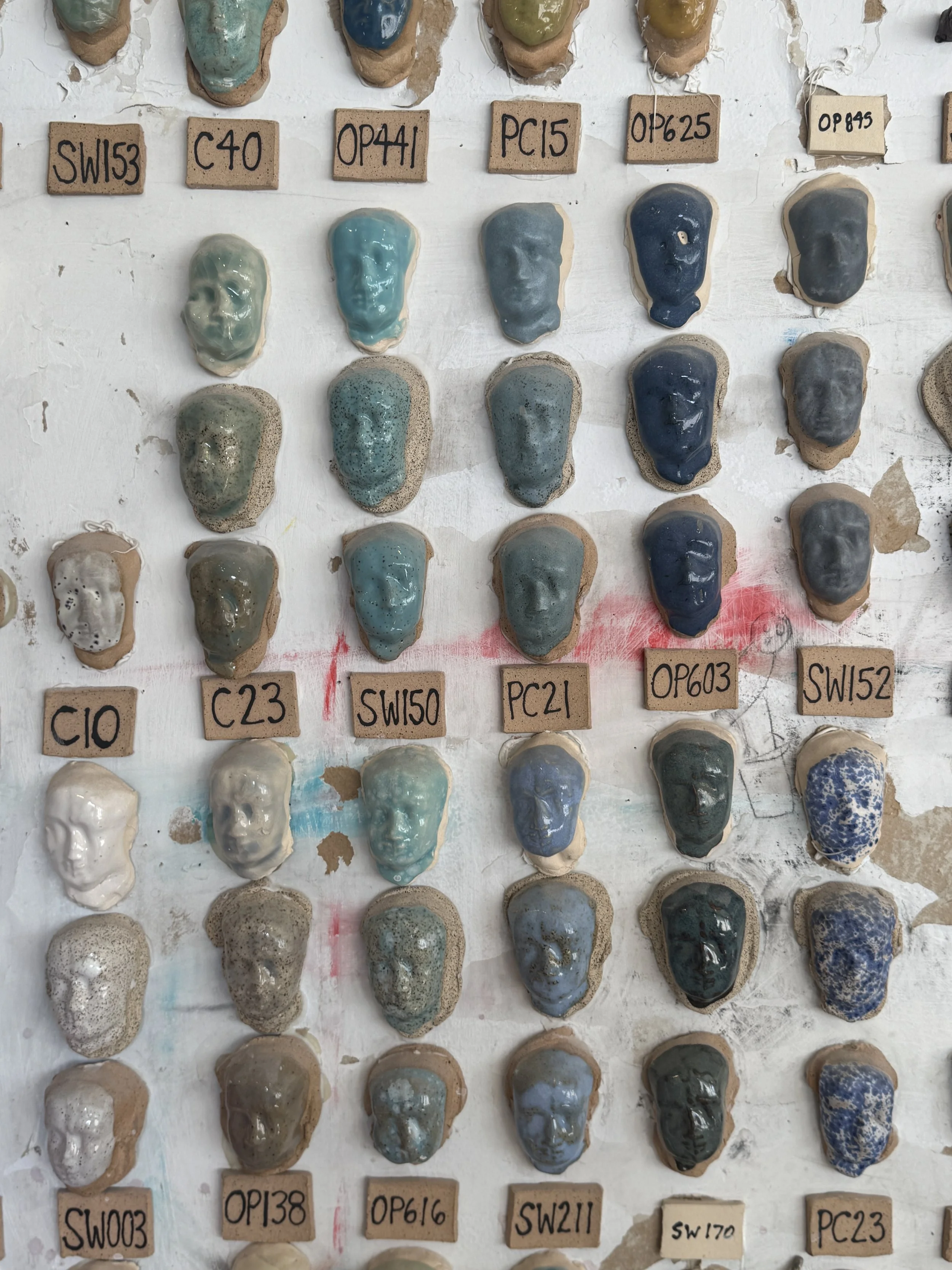 Close-up of a collection of ceramic slips or glazes in various shades of blue, green, gray, and speckled patterns, each on small clay bases with handwritten labels underneath.