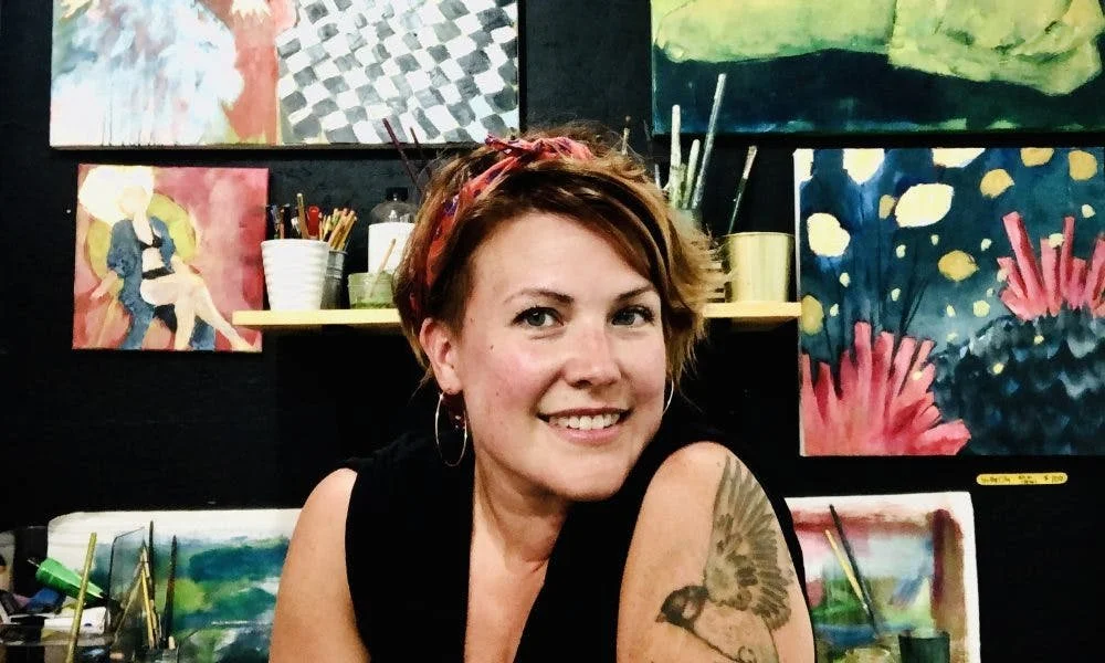 A woman with short brown hair, wearing hoop earrings and a headband, smiling in front of colorful abstract paintings in an art studio.