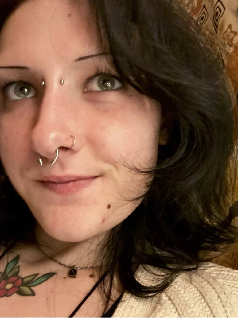 Close-up of a woman with black wavy hair, wearing multiple facial piercings including a septum ring, nostril piercings, and eyebrow piercings. She has a tattoo of flowers on her chest and is wearing a necklace with a small pendant. The background shows a patterned fabric or curtain.