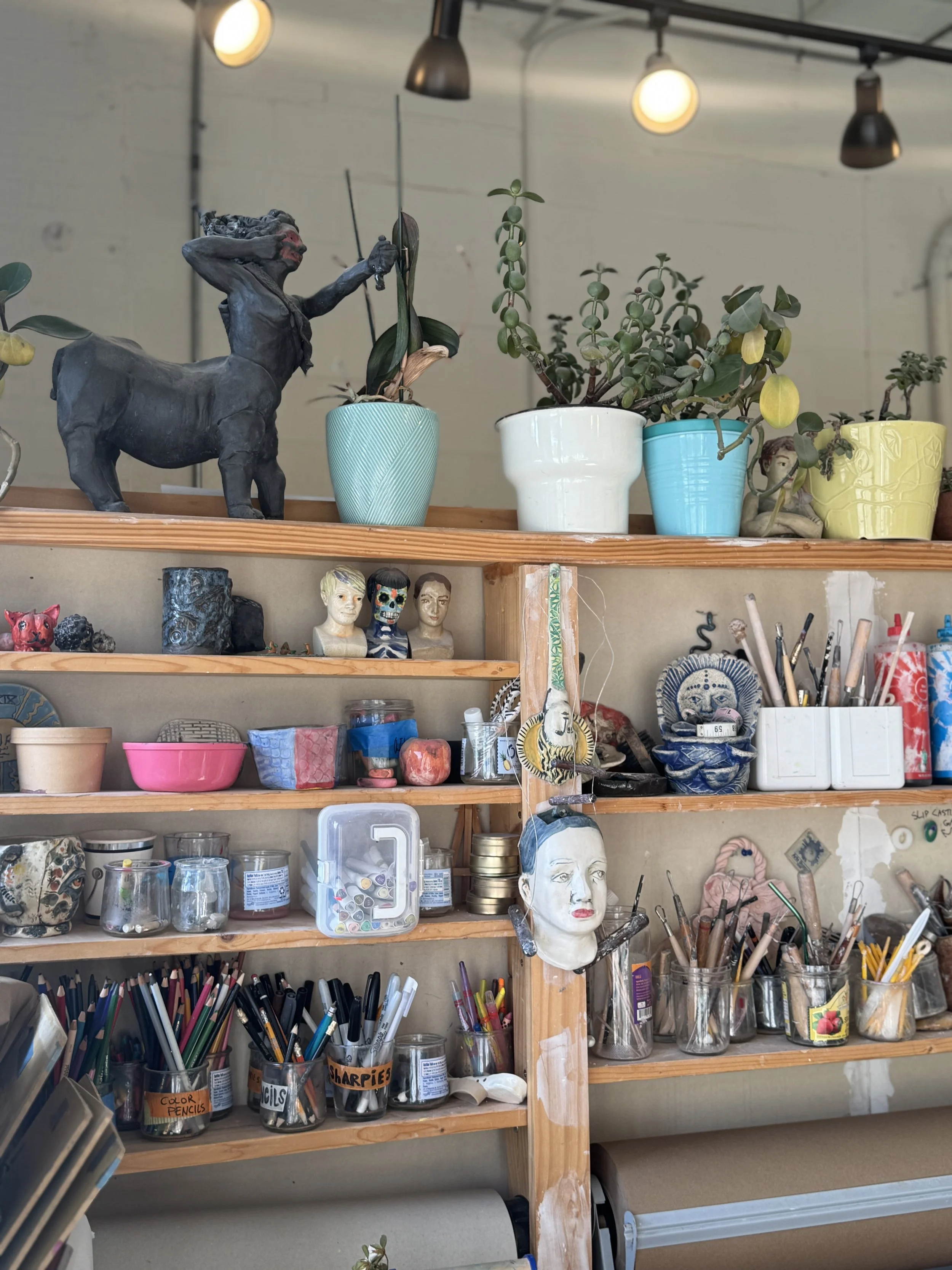 A wooden shelf filled with various art supplies including potted plants, sculpture, jars with pens and markers, and decorative objects, with overhead lights illuminating the space.