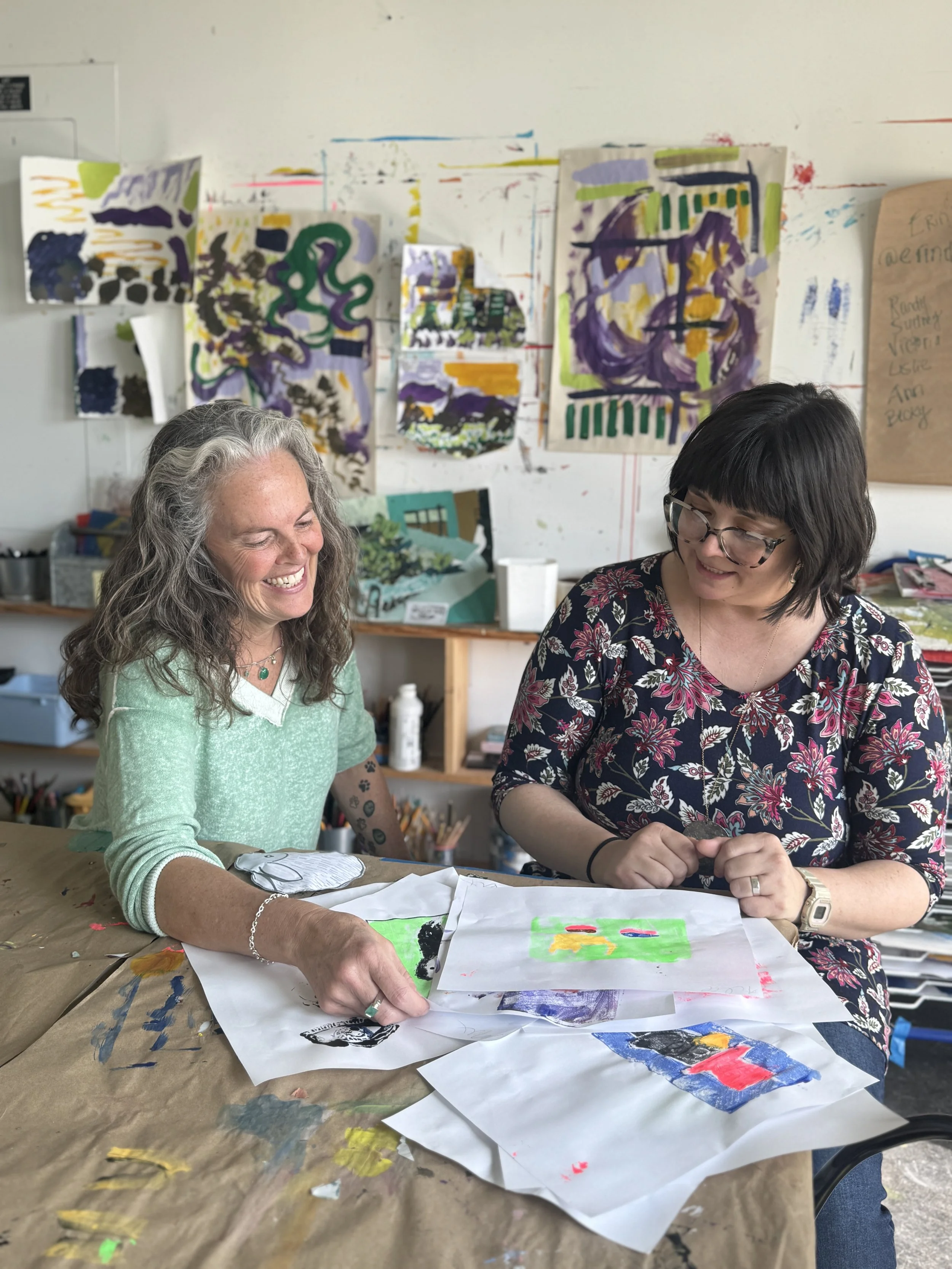 Two women with dark hair and glasses, smiling and working on art projects at a wooden table covered with papers and paint. Artworks are displayed on a wall behind them.