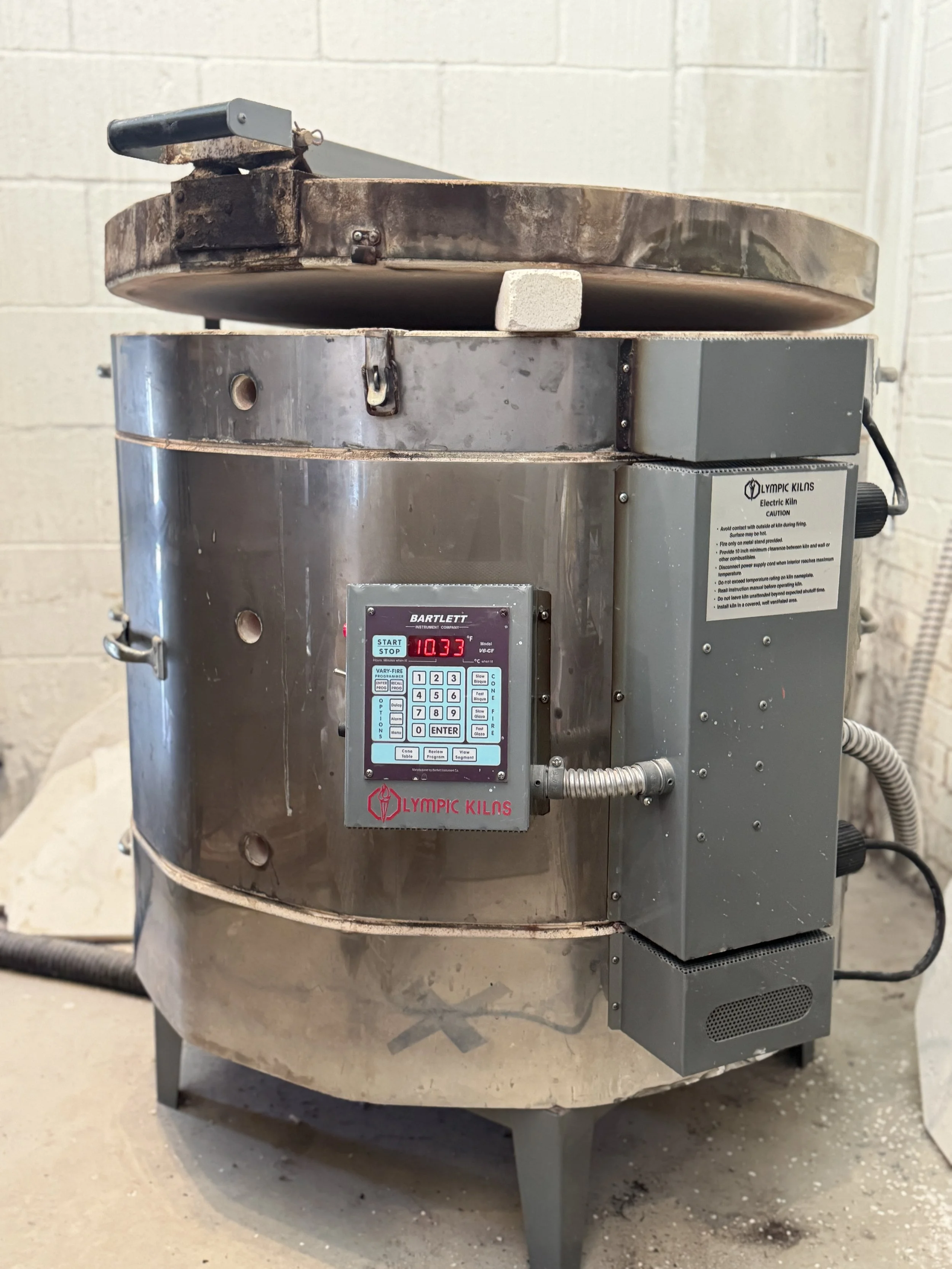 Industrial electric kiln with digital display showing temperature, metal exterior, and electrical control panel, labeled 'Olympic Kilns' in a workshop setting.