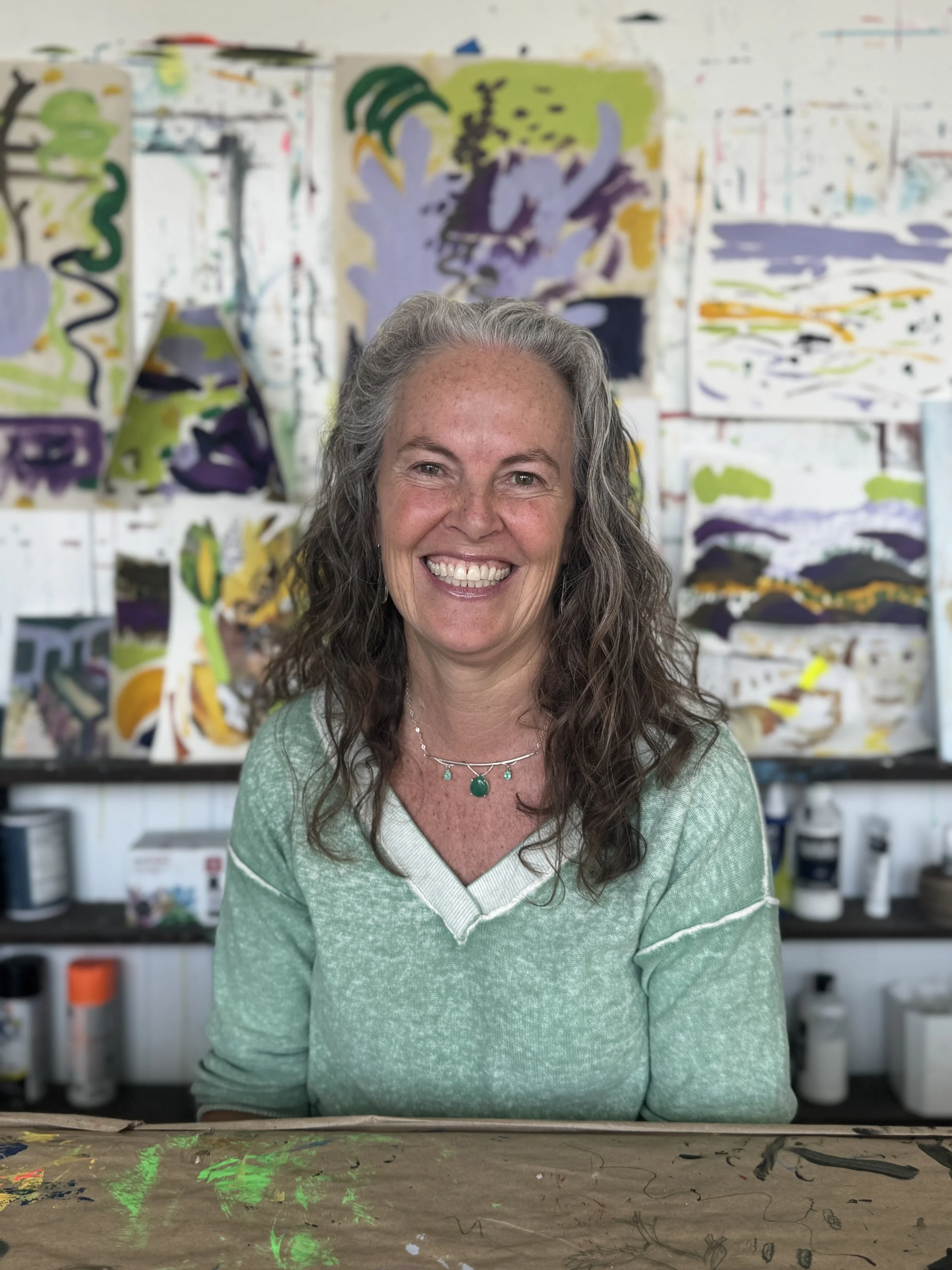 A smiling woman with gray hair, wearing a green sweater and a green necklace, sitting in front of colorful abstract paintings in an art studio.