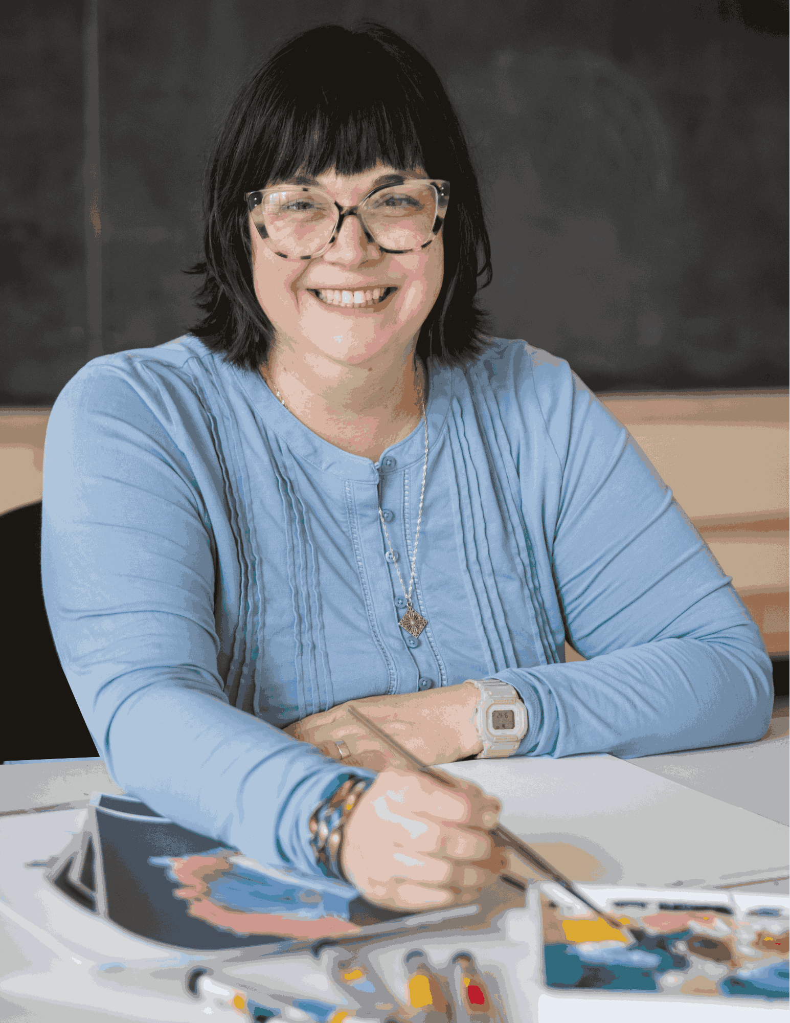 A woman with shoulder-length black hair wearing glasses and a light blue blouse, smiling while sitting at a table with art supplies, including paints and brushes.