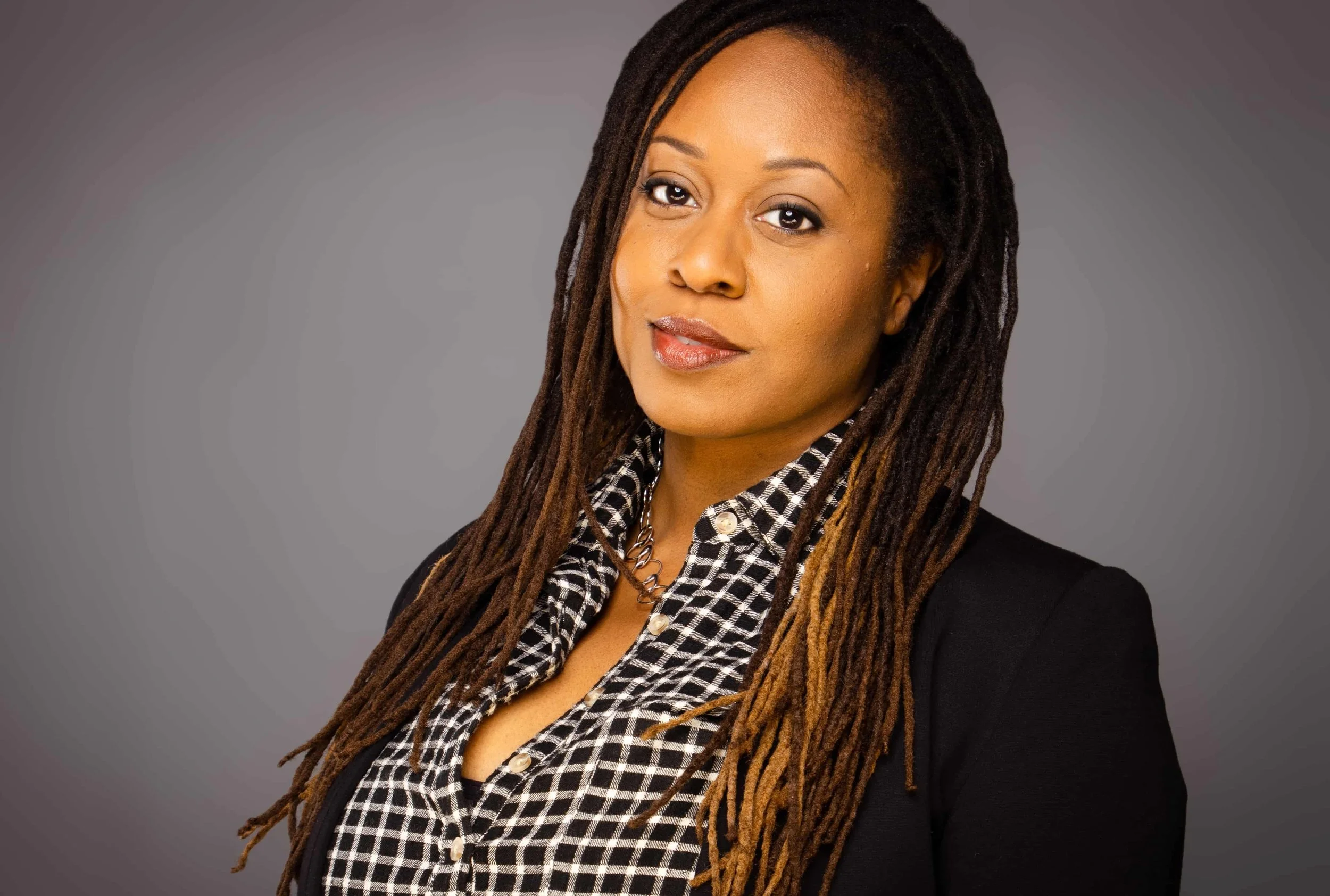 A woman with long dreadlocks wearing a black blazer and a black-and-white checkered shirt, looking at the camera with a slight smile, against a gray background.