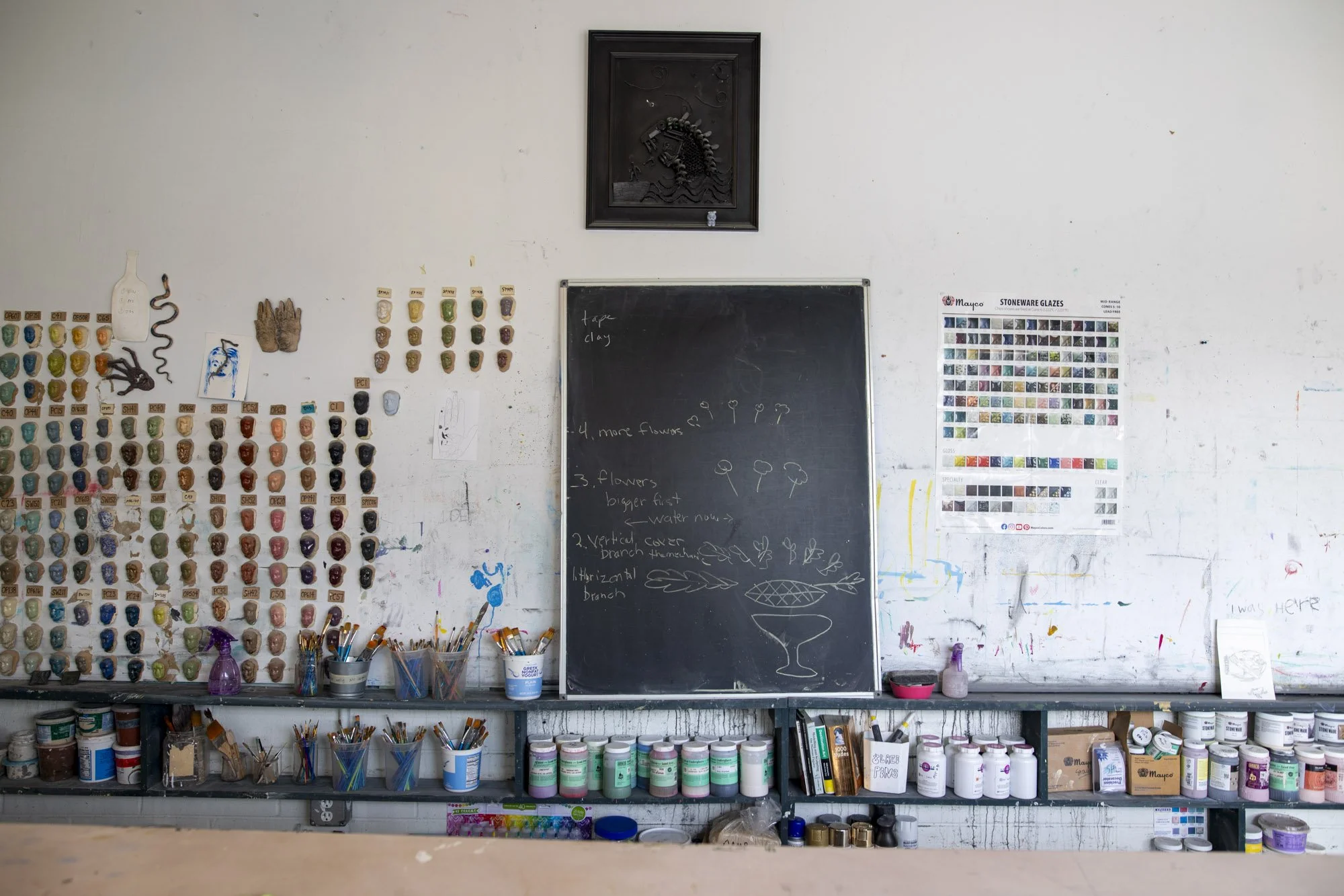 An art studio wall with various paint colors, brushes, and supplies, a black chalkboard with handwritten notes, and a framed black sculpture of a horse's head.